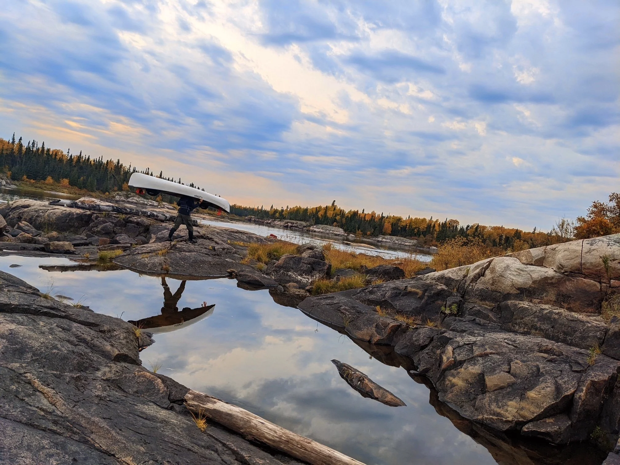 A person carrying a white kayak over their shoulders walking on rocky terrain beside a river with a forested background under a partly cloudy sky.