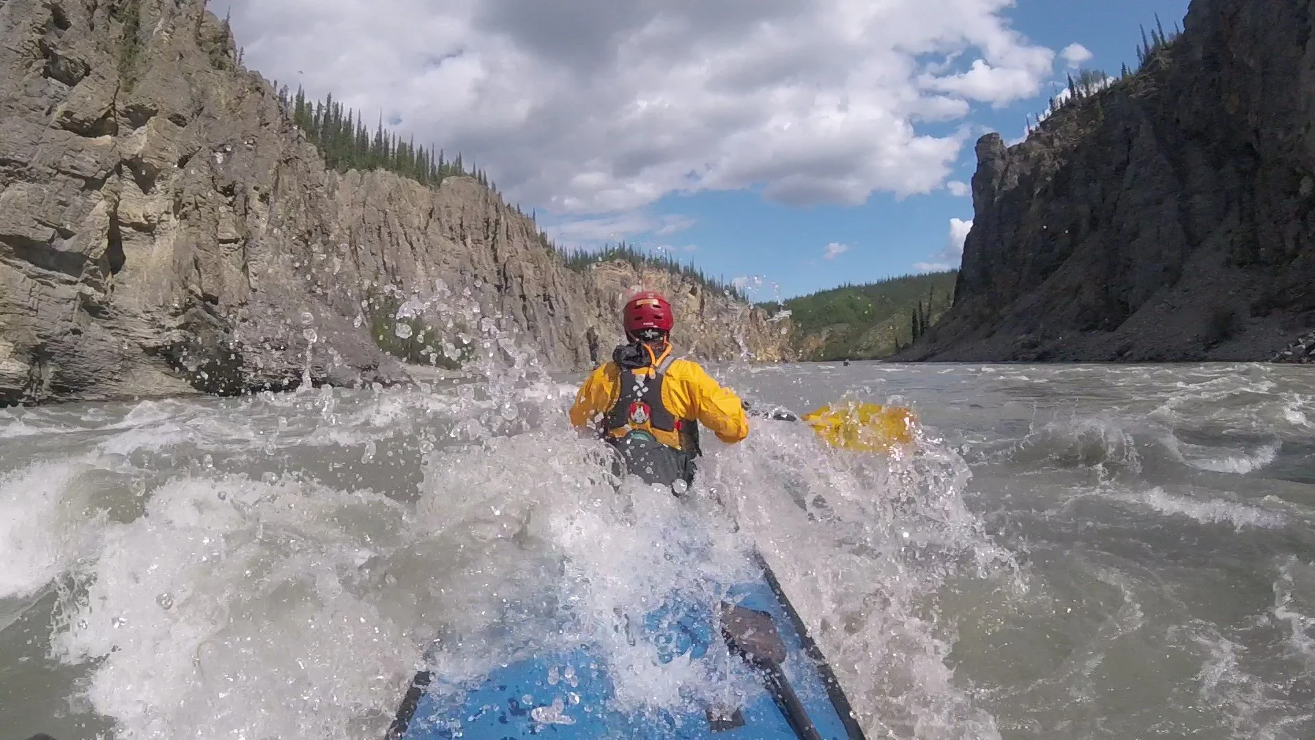 Person in yellow raft navigating rough river water with canyon cliffs and cloudy sky in the background.