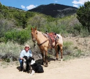woman kneeling with two border collies in front of her and saddled red dun horse beside her, background brush and trees in foothills