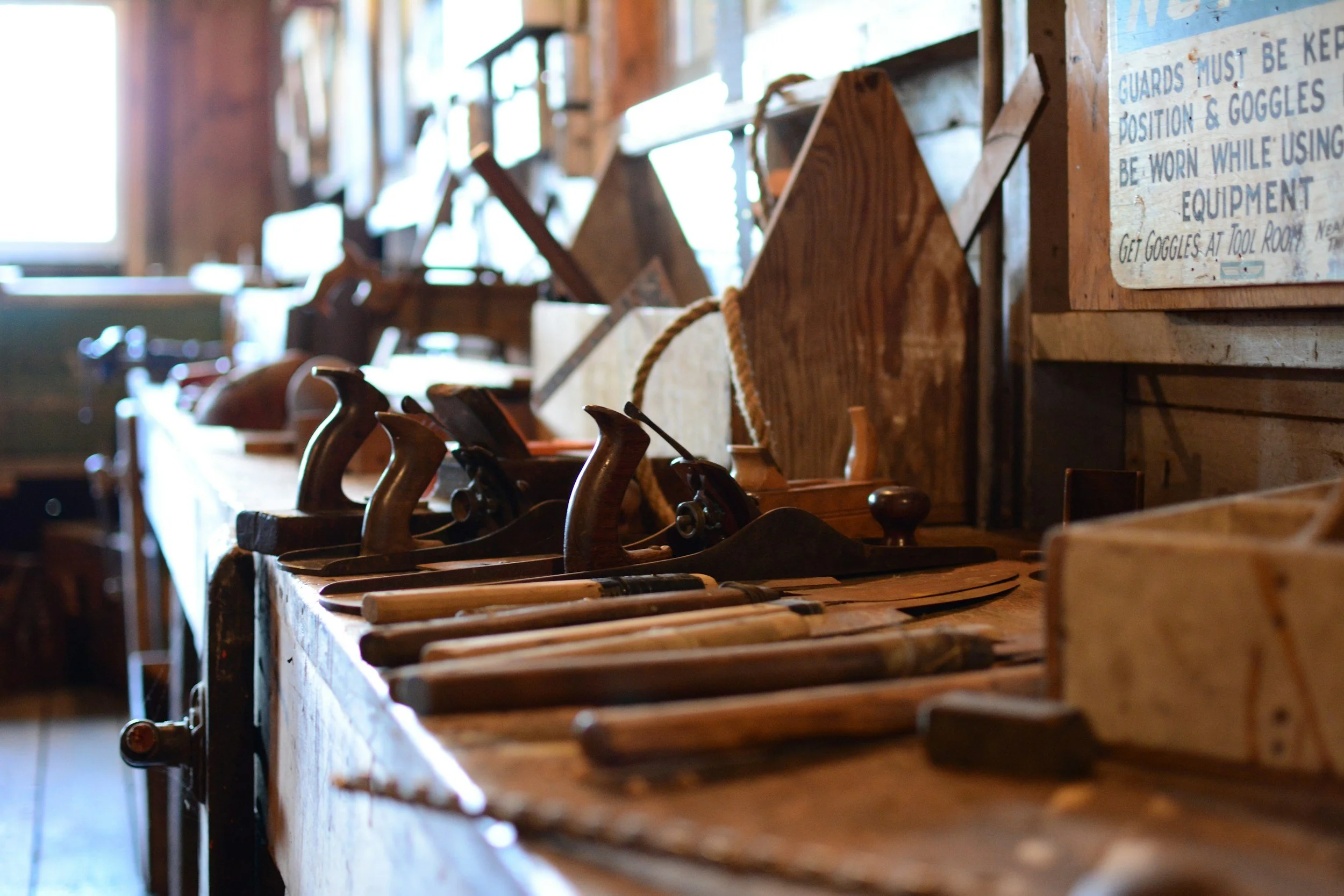 A workbench with vintage woodworking hand tools, including saws and drills, in a rustic workshop with wooden walls and a sign with safety instructions.