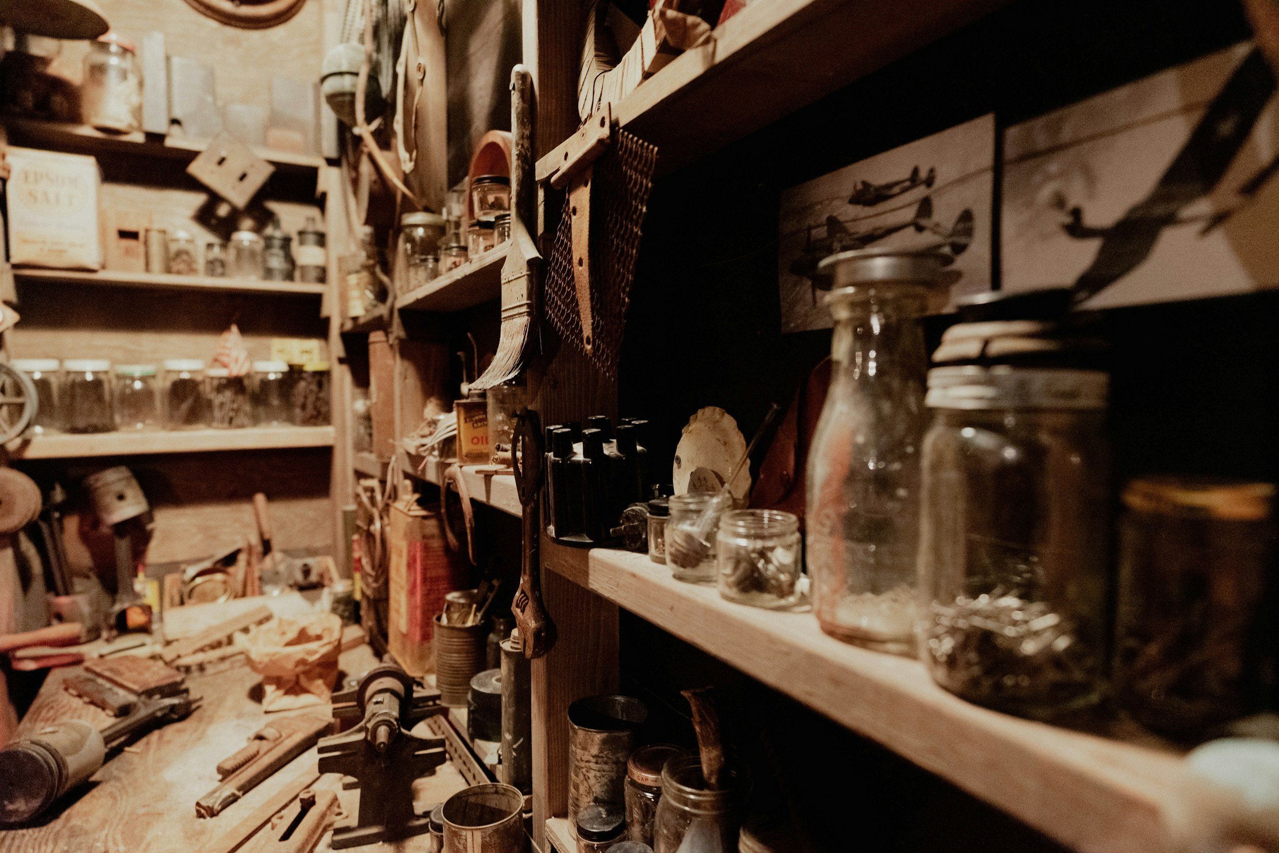 A cluttered workshop with wooden shelves filled with jars, tools, and various objects, and workbenches with drills and miscellaneous items.