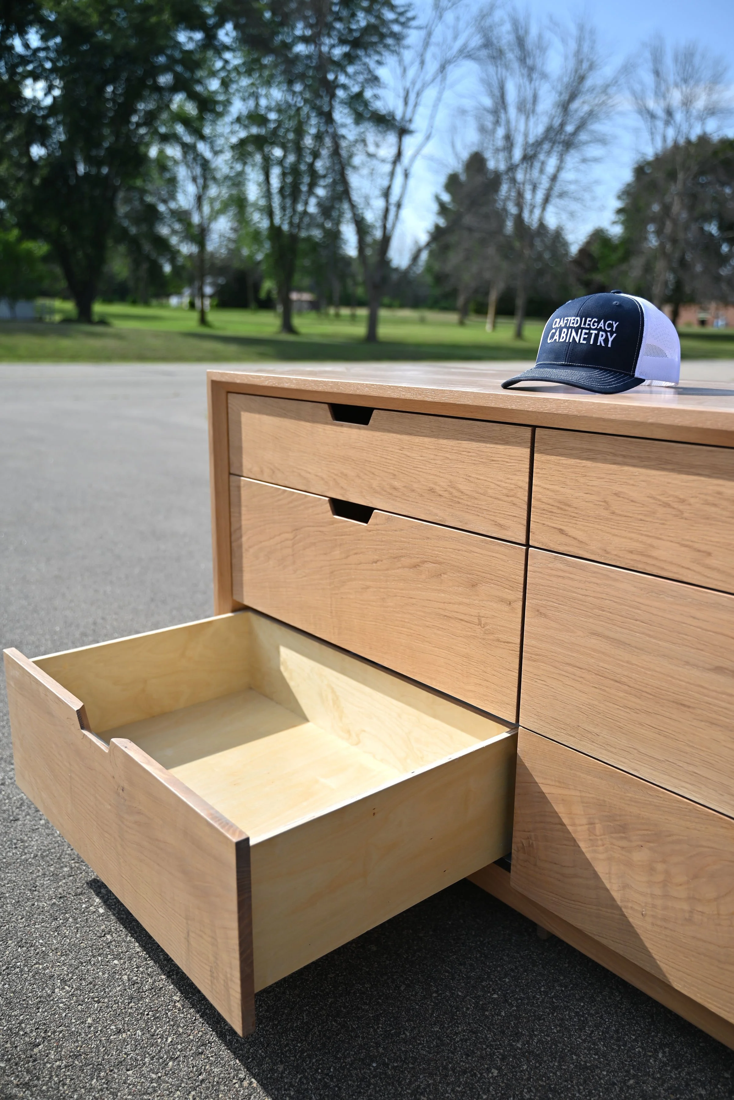 A wooden dresser with multiple drawers, one of which is pulled open, and a navy blue and white baseball cap on top that reads "Crafted Legacy Cabinetry." Mid century modern dresser . Avocado inspired 
Minimalist dresser   Minimalist furniture