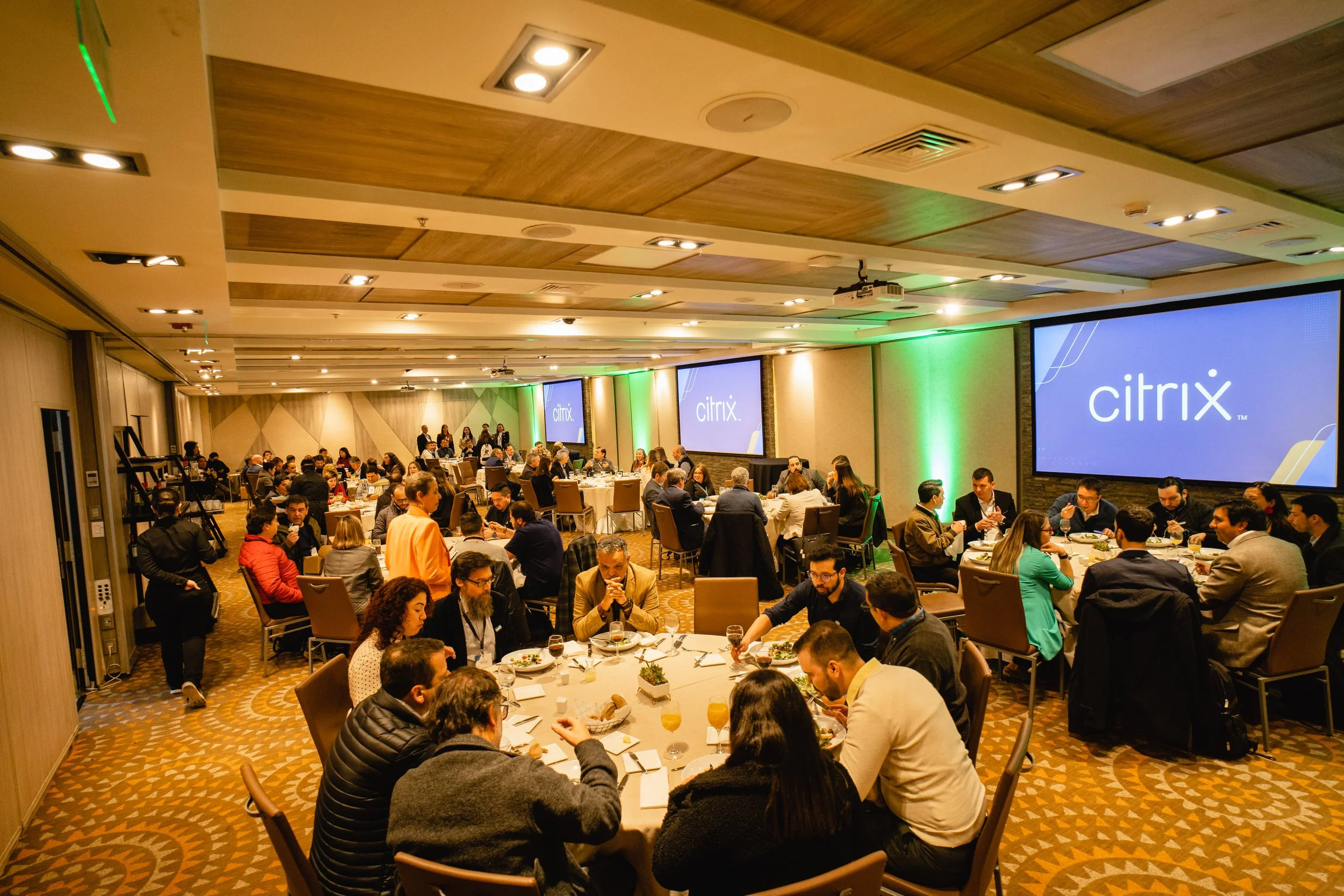 Conferencia o evento corporativo con muchas personas sentadas en mesas redondas y pantallas que muestran el logo de Citrix, en un salón con iluminación cálida y decoraciones en tonos verdes y beige.