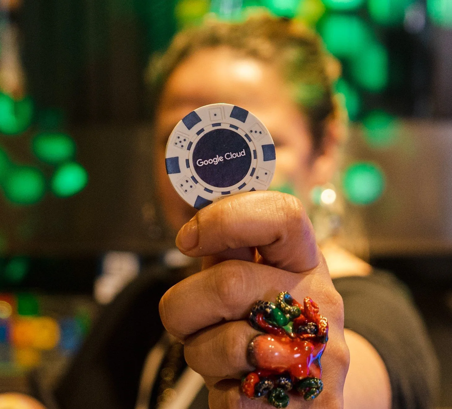 Persona sosteniendo una ficha de poker con el logo de Google Cloud, con fondo difuso de luces verdes y una persona al fondo.