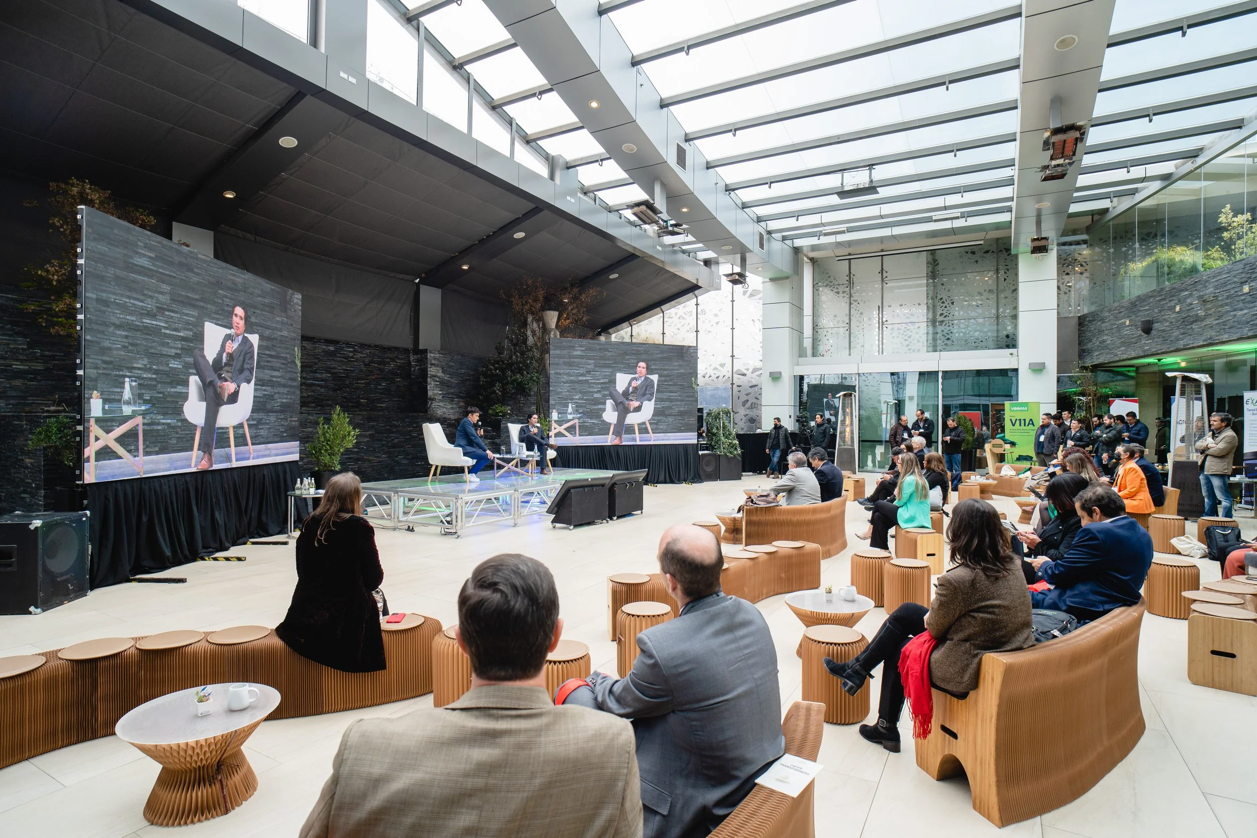 Auditorio moderno con asistentes en presencia y en línea, dos pantallas grandes muestran a un conferencista sentado en una silla blanca con micrófono, en un evento de conferencias o charla empresarial.