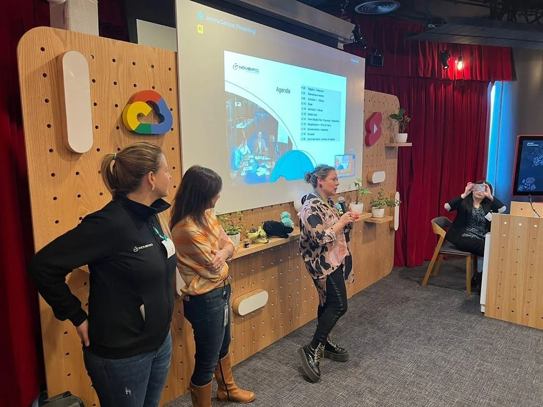 Tres mujeres presentan en un evento, una de ellas habla con un micrófono y las otras dos observan, mientras otra mujer toma una foto con su teléfono, en un entorno con paredes de madera y decoraciones, y un proyector con una diapositiva en la pantall