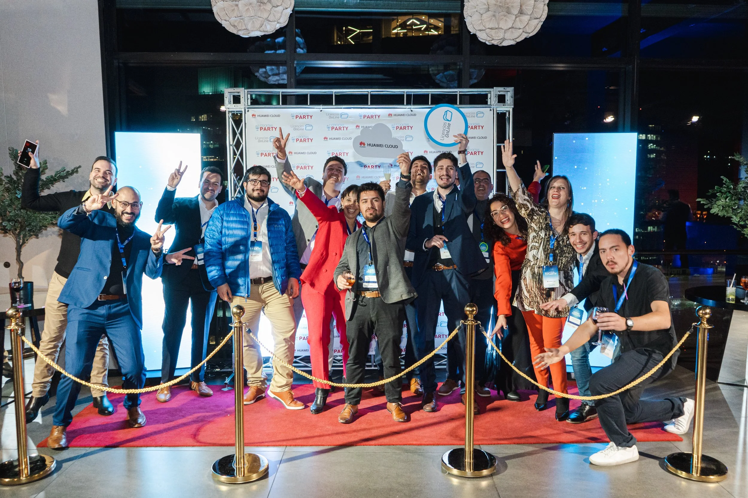 Grupo de personas en una celebración de evento tecnológico, posando frente a un fondo con logotipos de Huawei Cloud, algunos levantando la mano y haciendo la señal de paz, en un espacio con ambiente festivo y decoraciones modernas.