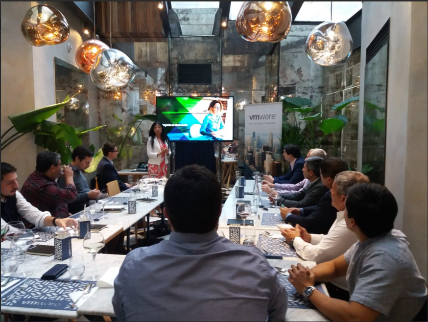 Reunión de negocios en un restaurante elegante, con participantes sentados en una mesa larga, una mujer presenta frente a una pantalla, ambiente moderno con plantas y luces decorativas.