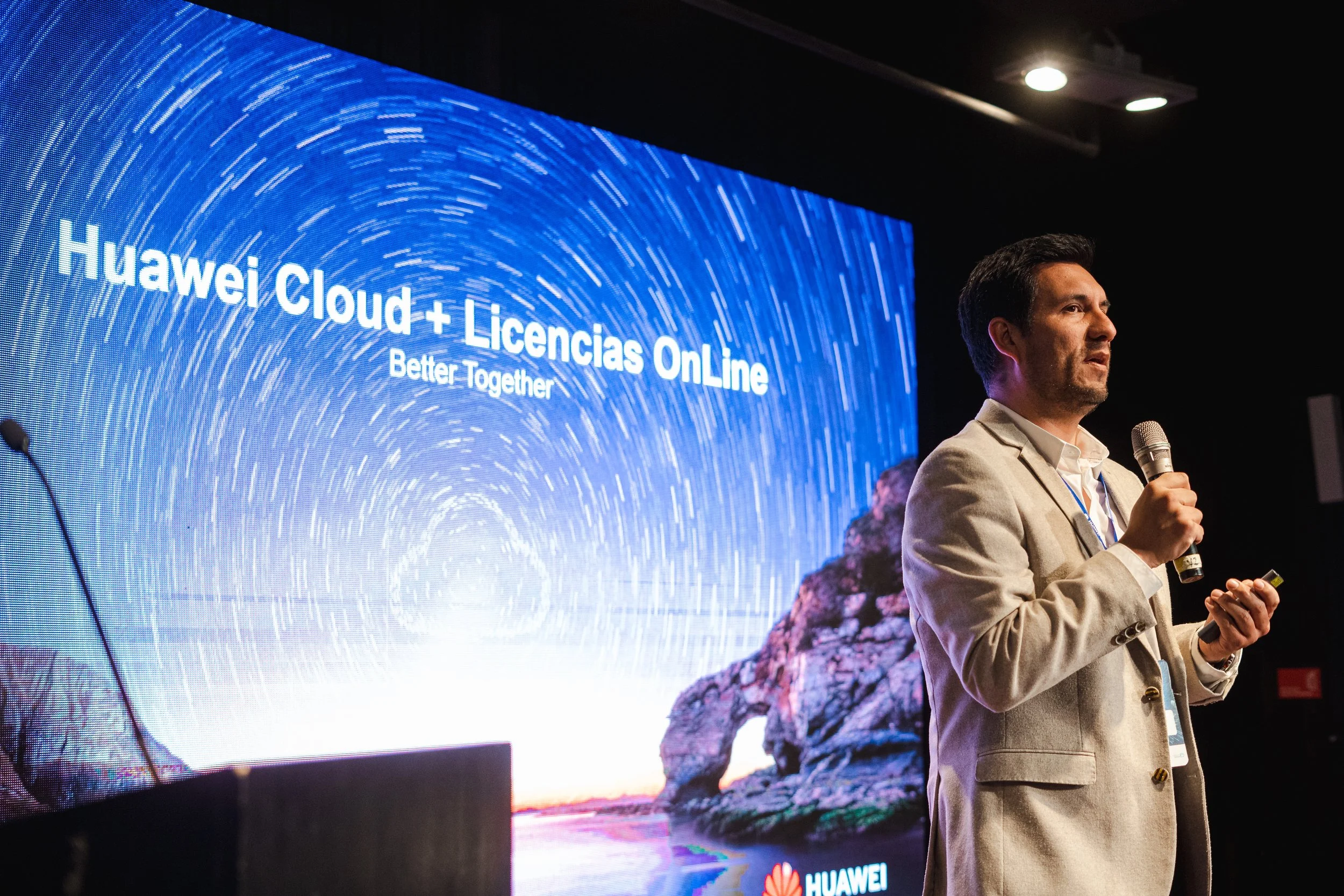 Hombre con saco claro y camisa blanca, sosteniendo un micrófono y una presentación en una conferencia sobre Huawei Cloud + Licencias OnLine.