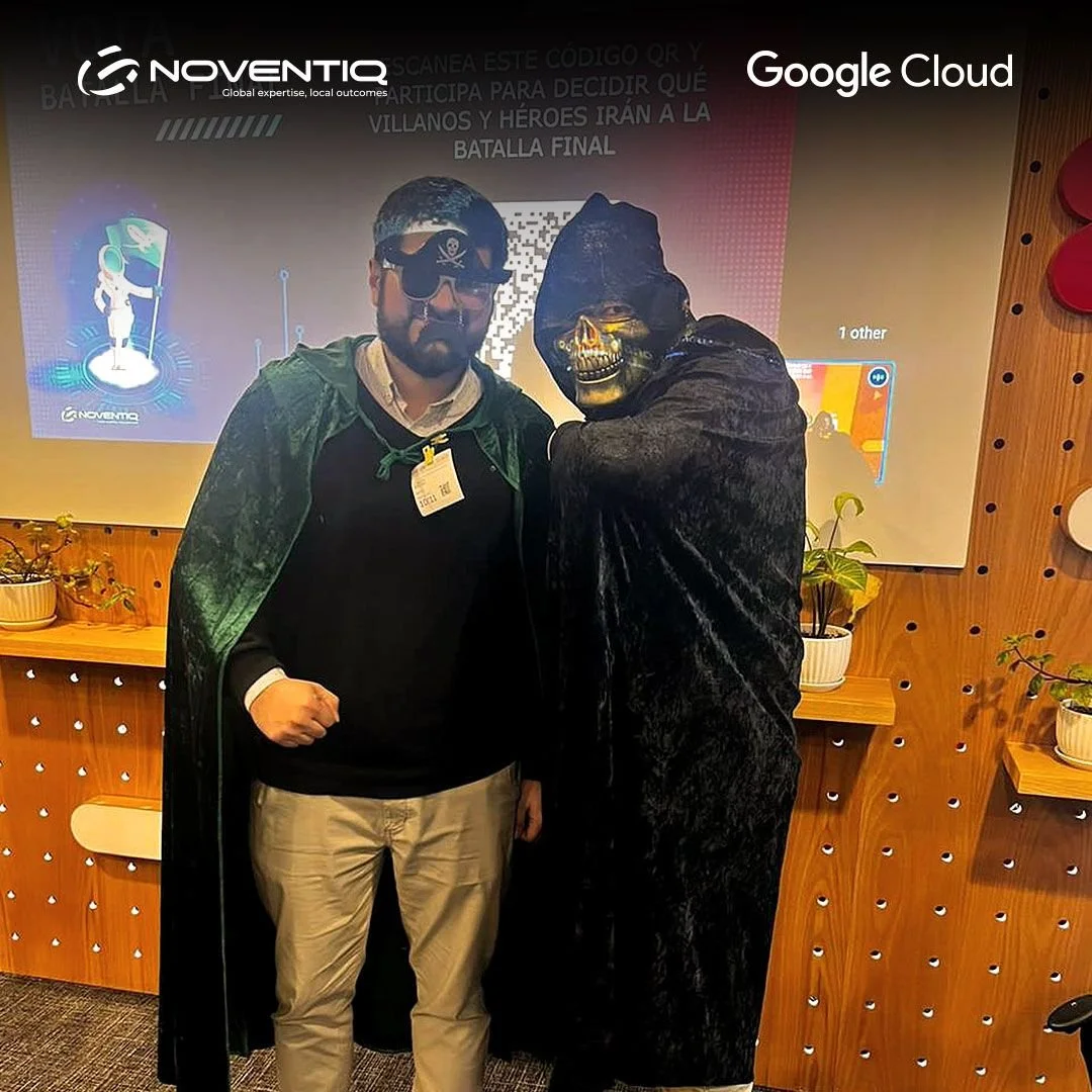 Dos personas disfrazadas, una con un disfraz de pirata y otra con un disfraz de calavera, posando juntos frente a una pantalla en un entorno de oficina o evento.