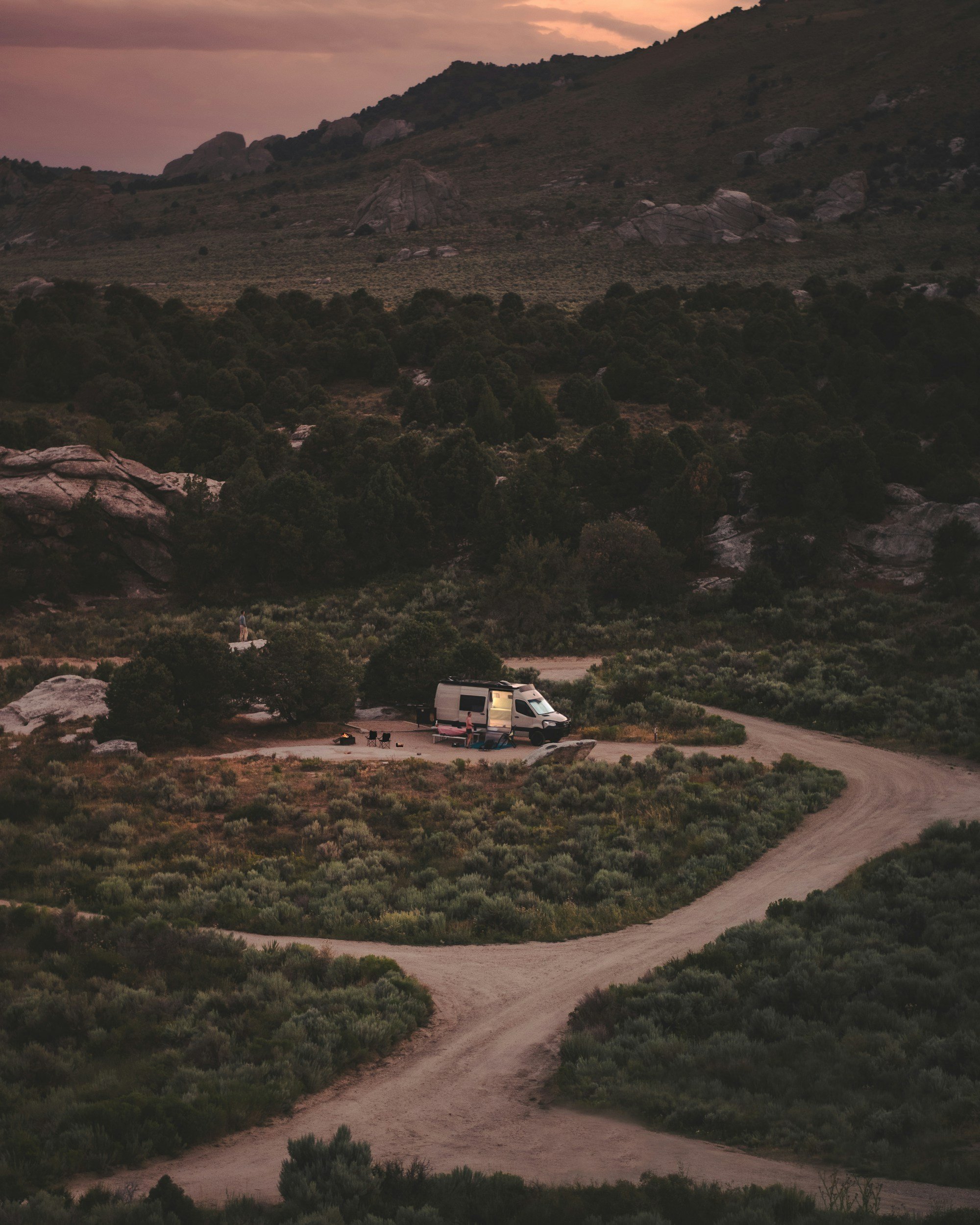 Campground with a white camper van and camping chairs on a winding dirt road in a mountainous area during sunset, surrounded by green bushes and trees.