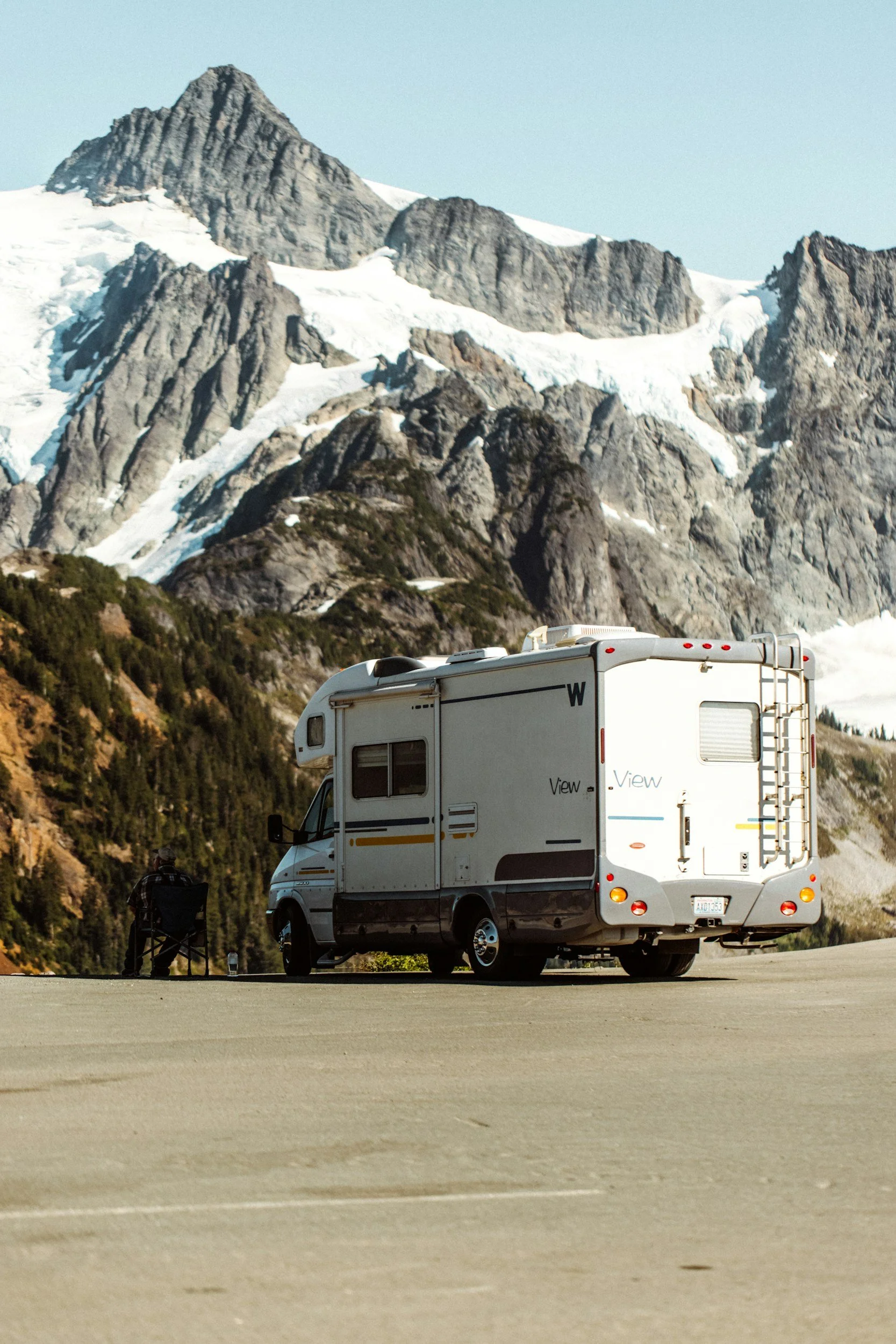 A white RV parked on a paved lot with mountains and snow in the background.