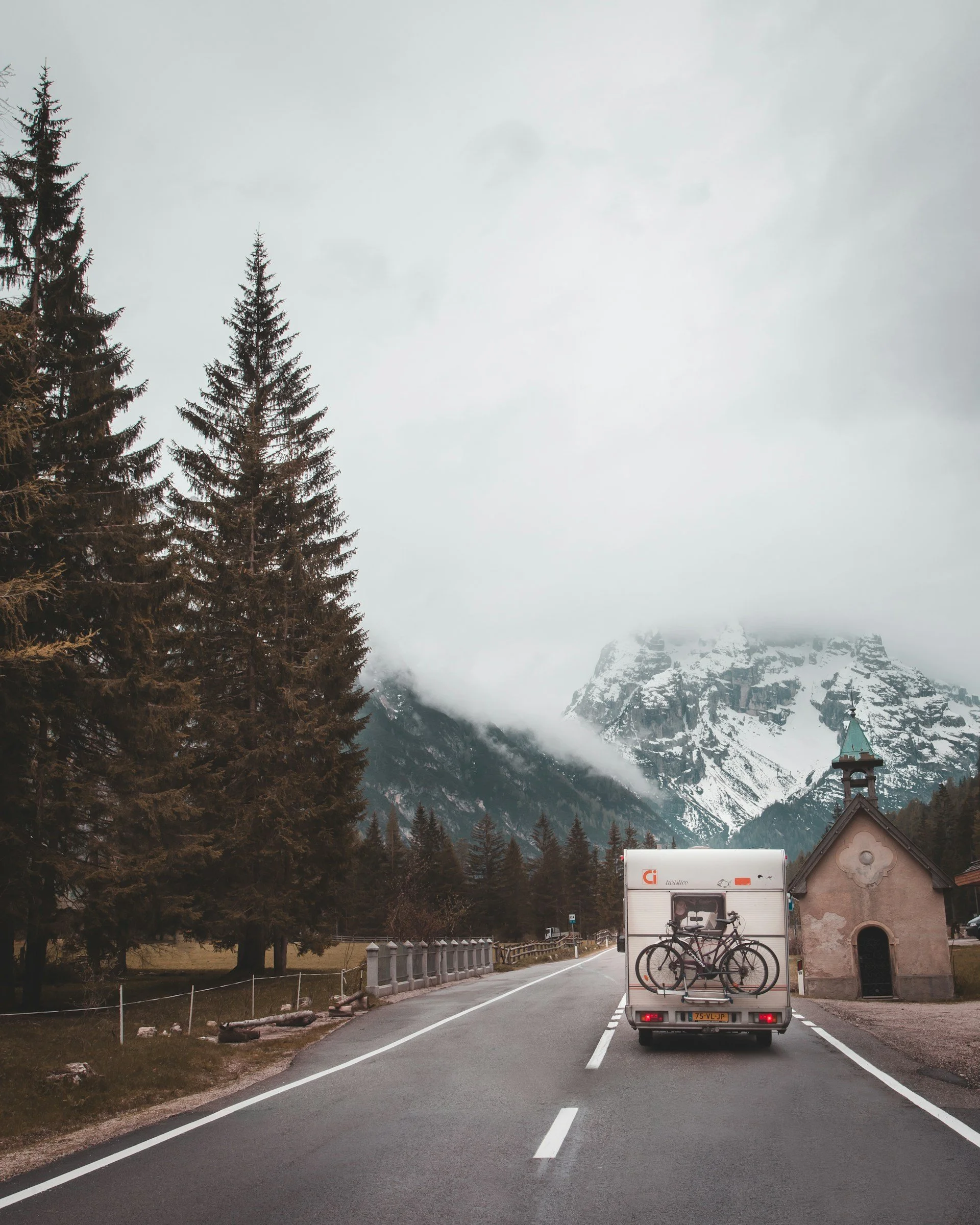 A small camper van with two bicycles attached to the back driving on a mountain road. The road is lined with a fence and has tall trees on one side and snow-capped mountains in the background. The sky is overcast.