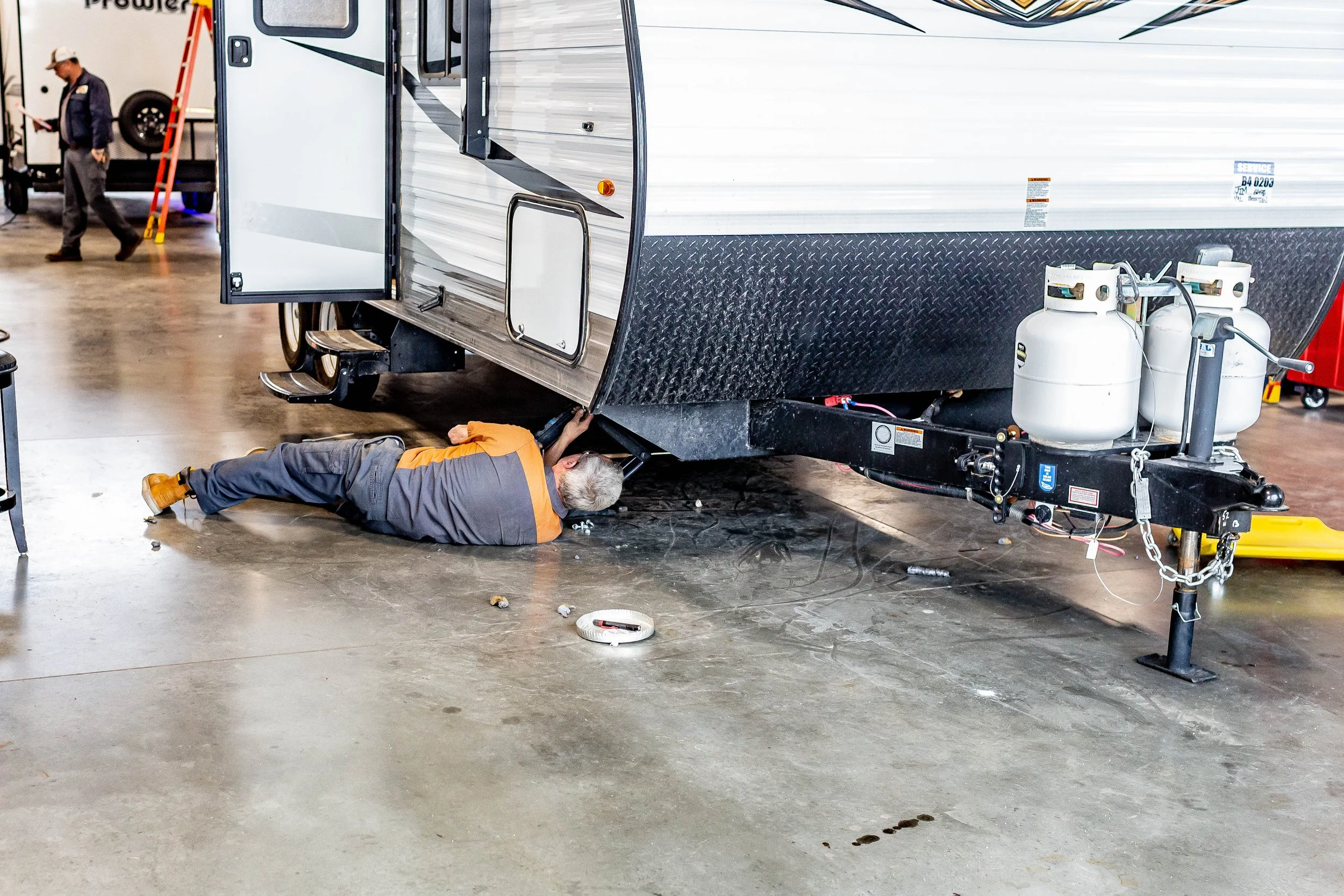 A person working underneath a travel trailer or RV inside a large indoor facility.