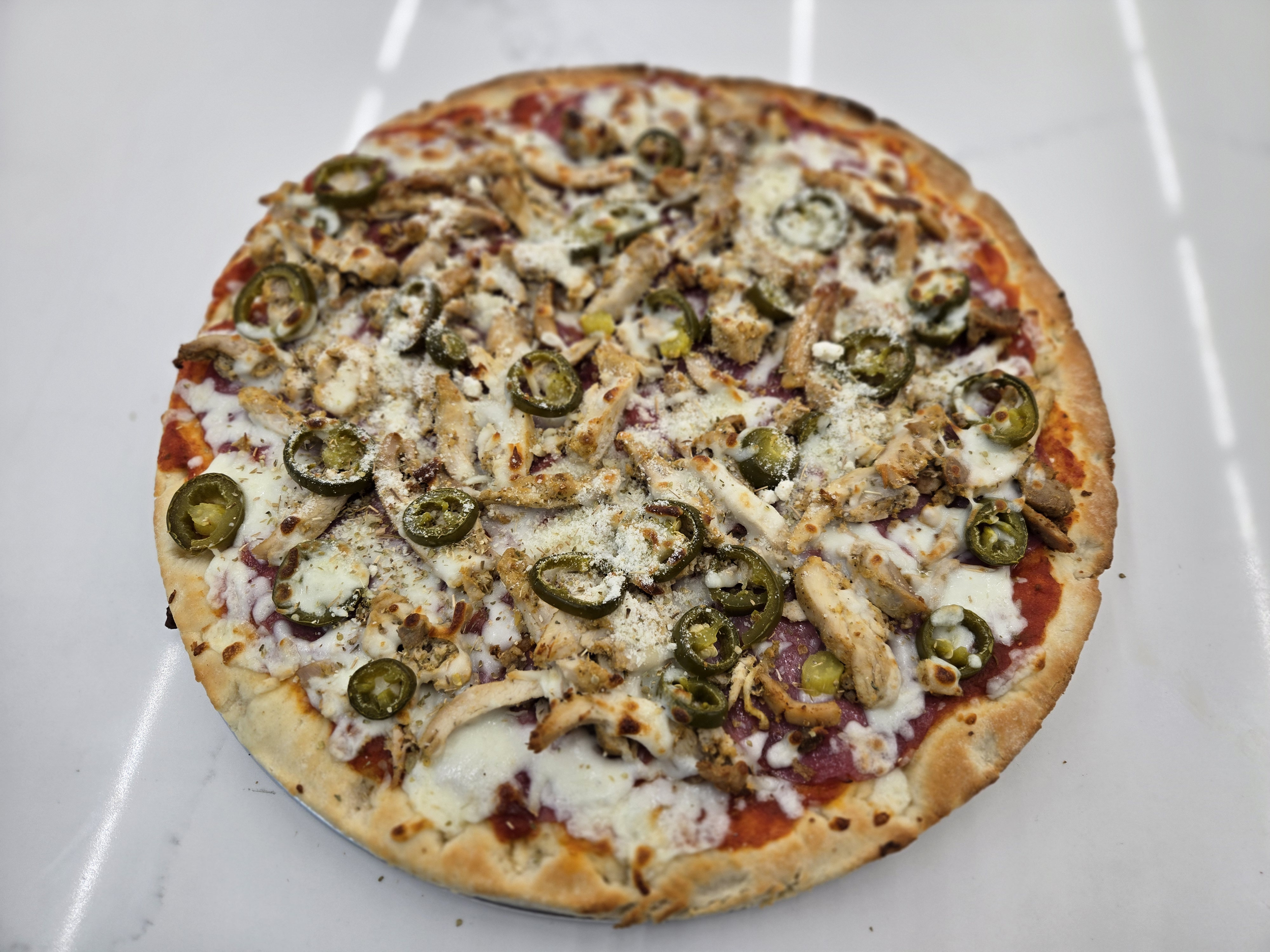 A loaded pizza with cheese, sliced jalapeños, shredded chicken, and other toppings on a white surface.