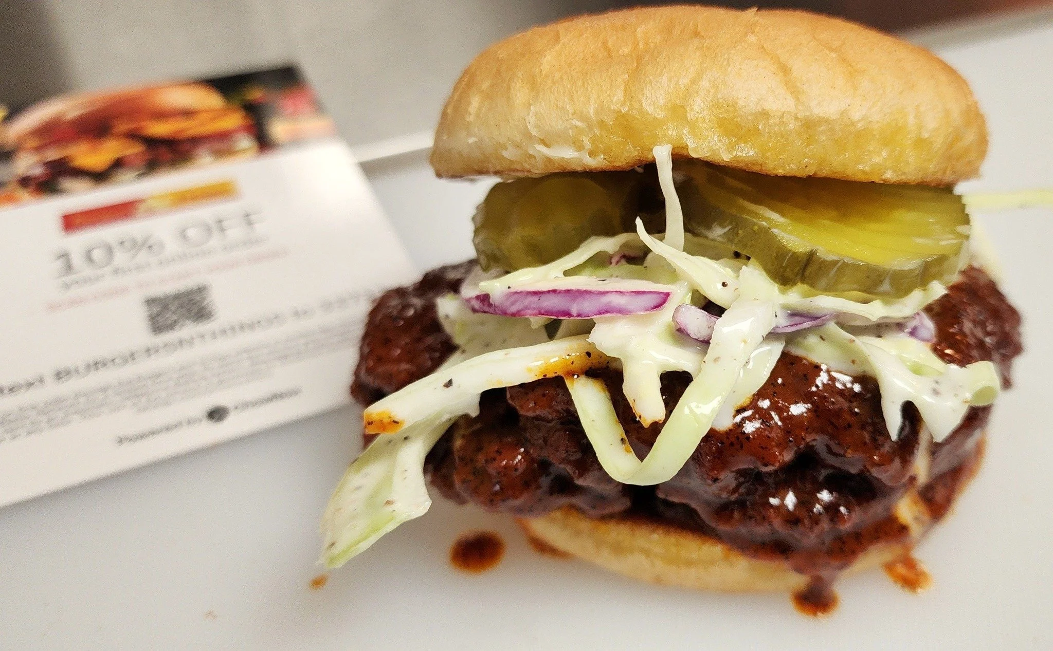 A burger with a beef patty, coleslaw, and pickles in a bun on a white surface.