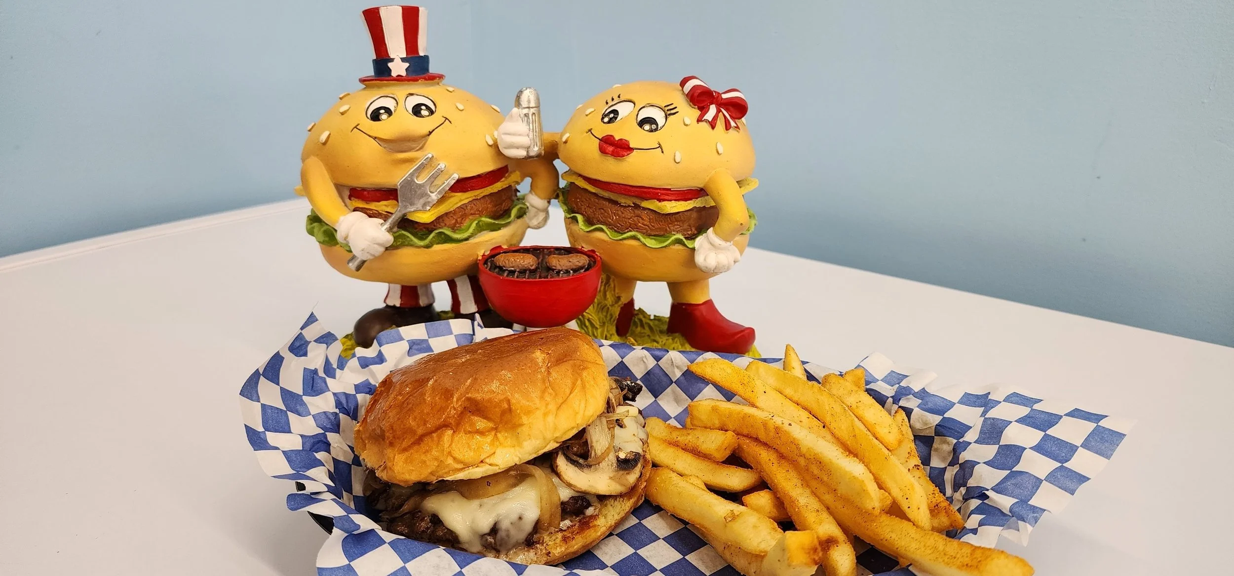 A cheeseburger with mushrooms and melted cheese, served with French fries, on a checkered paper, with decorative burger-themed figurines and a grill in the background.