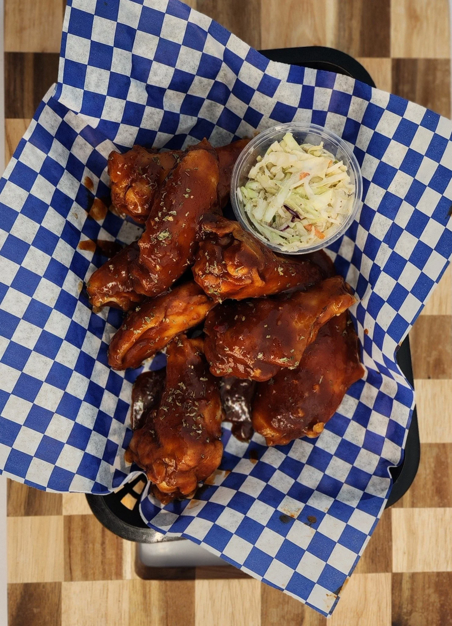 Basket of buffalo chicken wings with buffalo sauce, served with a side of coleslaw in a small cup, on a checkered paper liner.