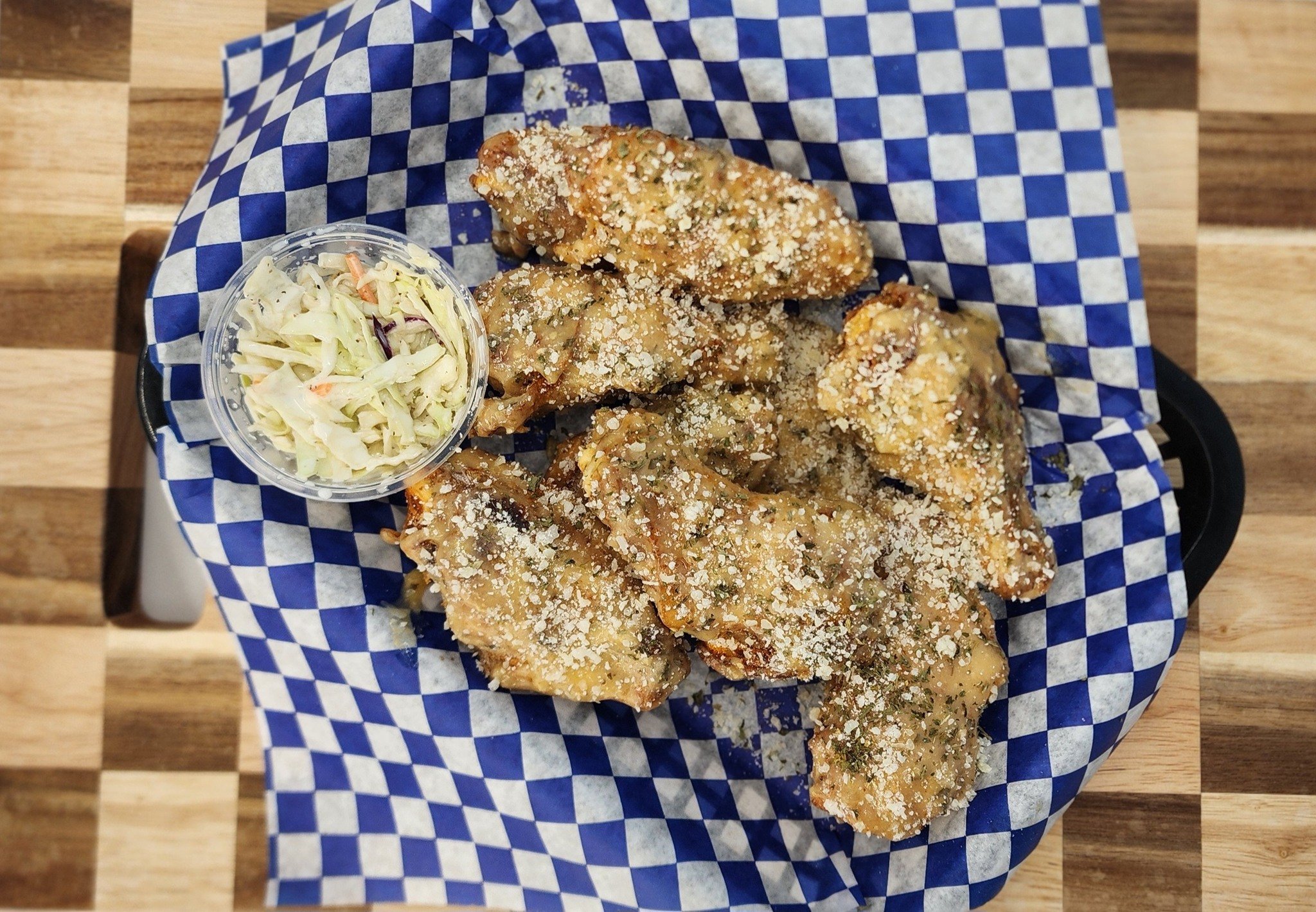 Fried chicken wings topped with grated cheese and herbs, served with a side of coleslaw in a small plastic container on a checkered blue and white paper liner.
