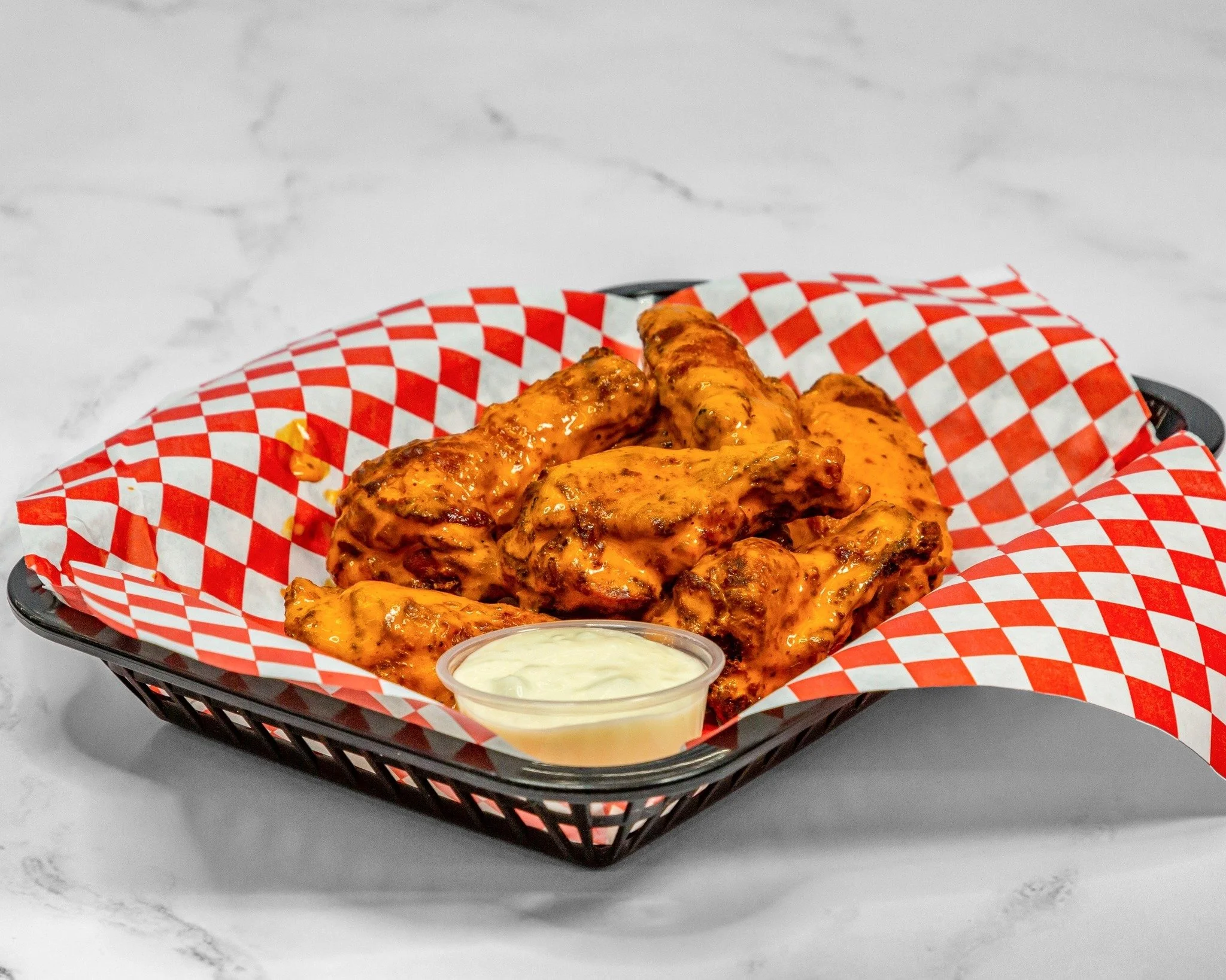 Basket of chicken wings with buffalo sauce and a side of ranch dipping sauce on checkered paper lining in a black basket on a white marble surface.