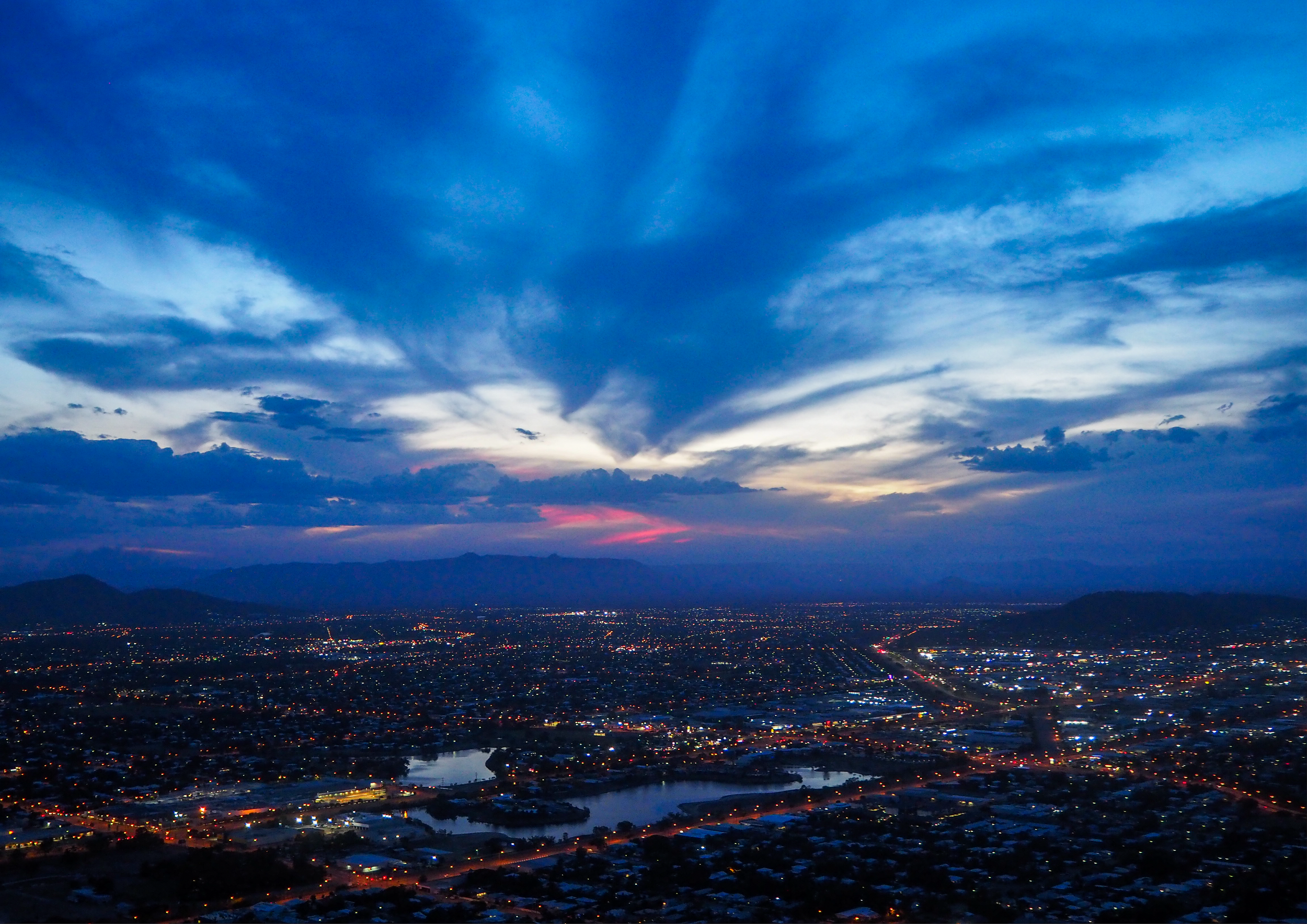 A panoramic view of a cityscape at dusk with a sky filled with blue clouds and a hint of sunset colors near the horizon. The city lights are beginning to turn on, illuminating the streets and buildings below.