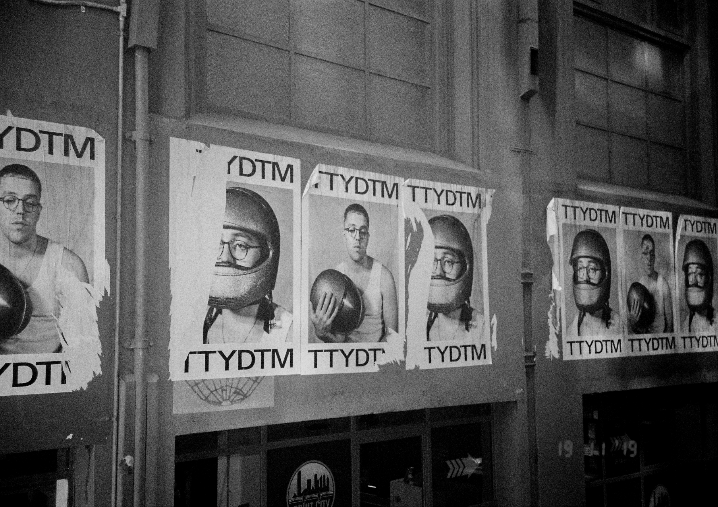 Black and white photograph of posters on a wall featuring multiple images of a young man wearing glasses and a helmet, with the text 'TYTTDTM' across the posters in an urban setting.