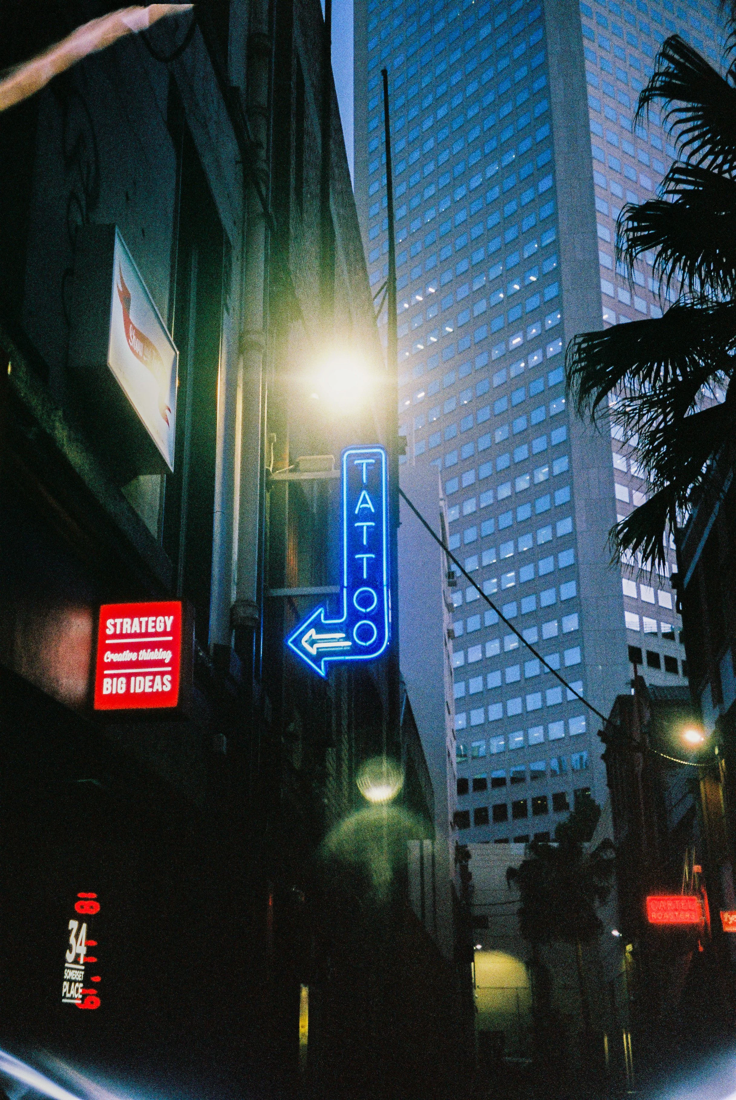 Nighttime city street with neon signs, including a blue arrow sign pointing left and a blue 'ATTENTION' sign, tall buildings, some palm trees, and streetlights.
