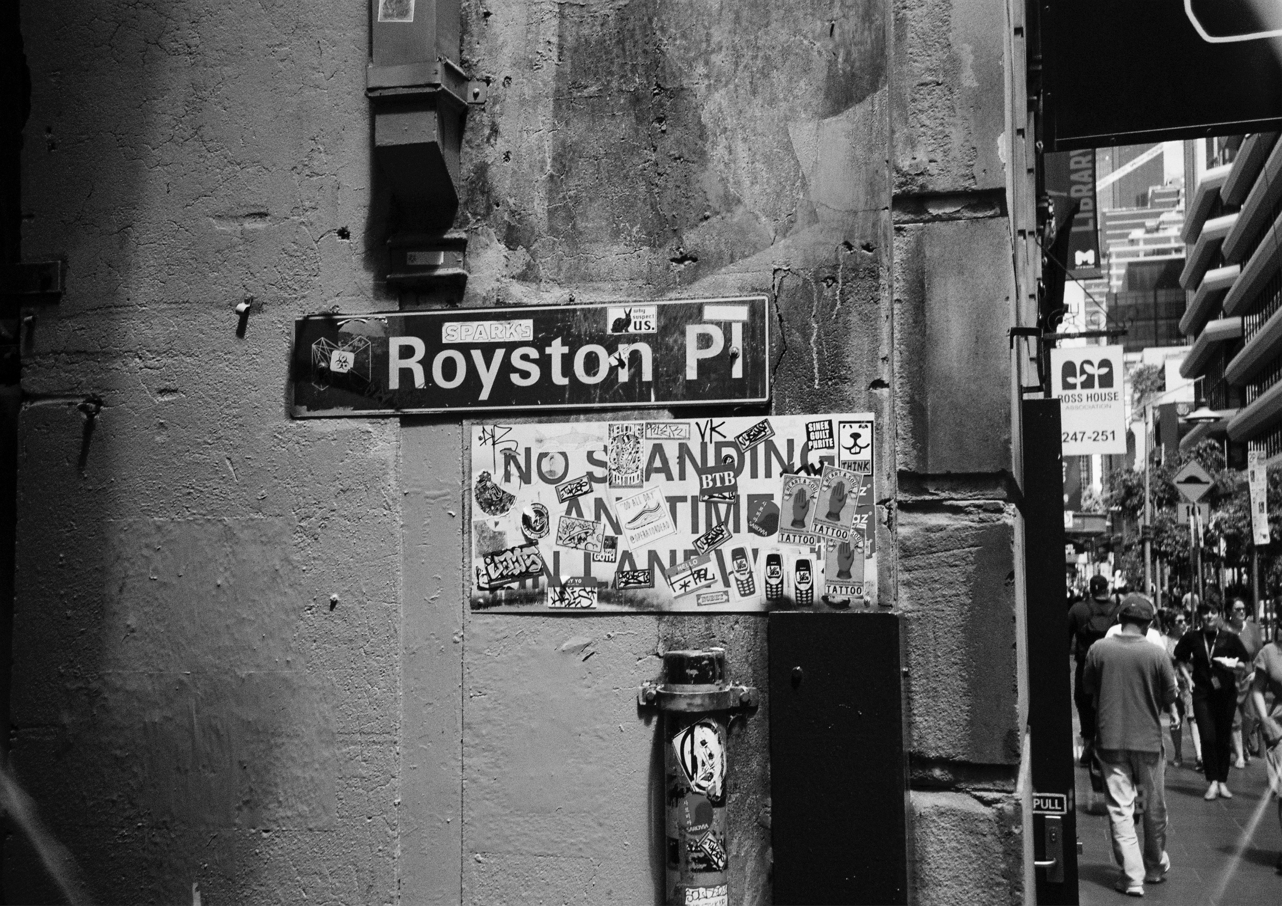 Melbourne street scene with a street sign that reads 'Royston P.', covered with stickers, and a group of people walking on the sidewalk. Black & White