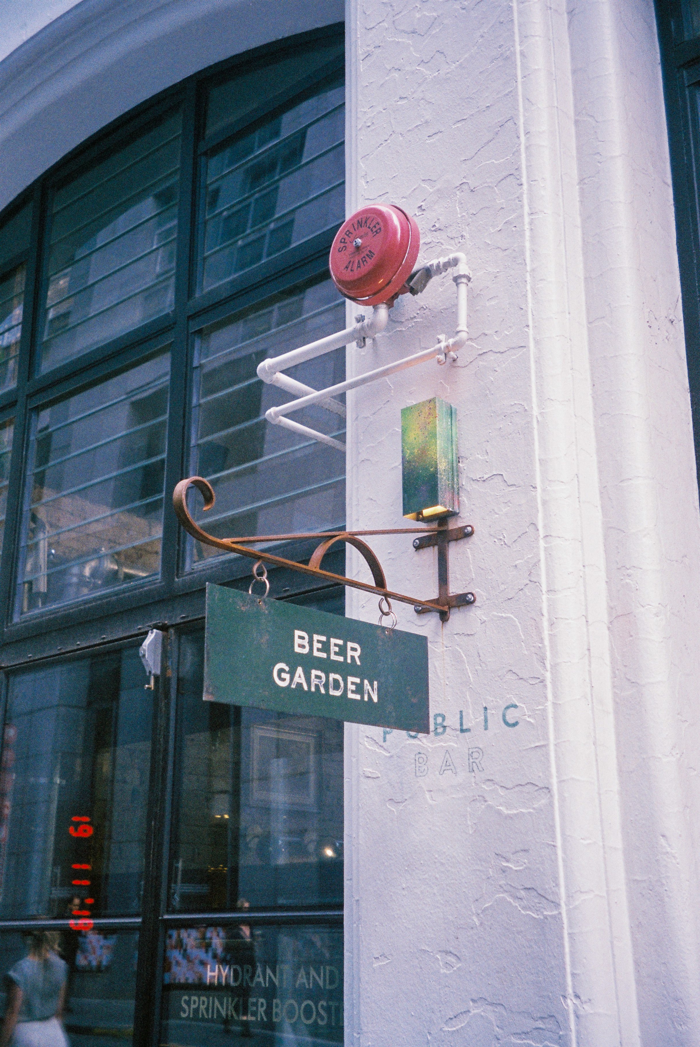 Exterior of a building with a sign that reads 'Beer Garden' hanging on a metal arm. There is a red fire alarm bell on the wall above the sign, and a small, multicolored rectangular box attached to the wall. The building has large dark-framed windows 