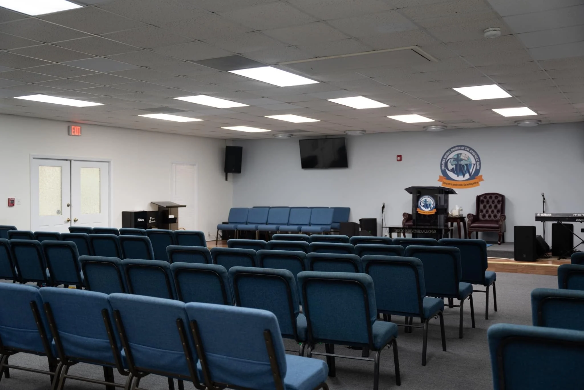 Empty church sanctuary with rows of blue chairs facing a stage that has a pulpit, a maroon chair, a piano, and a keyboard. There are two wall-mounted televisions and an emblem of the United Methodist Church on the wall behind the stage.