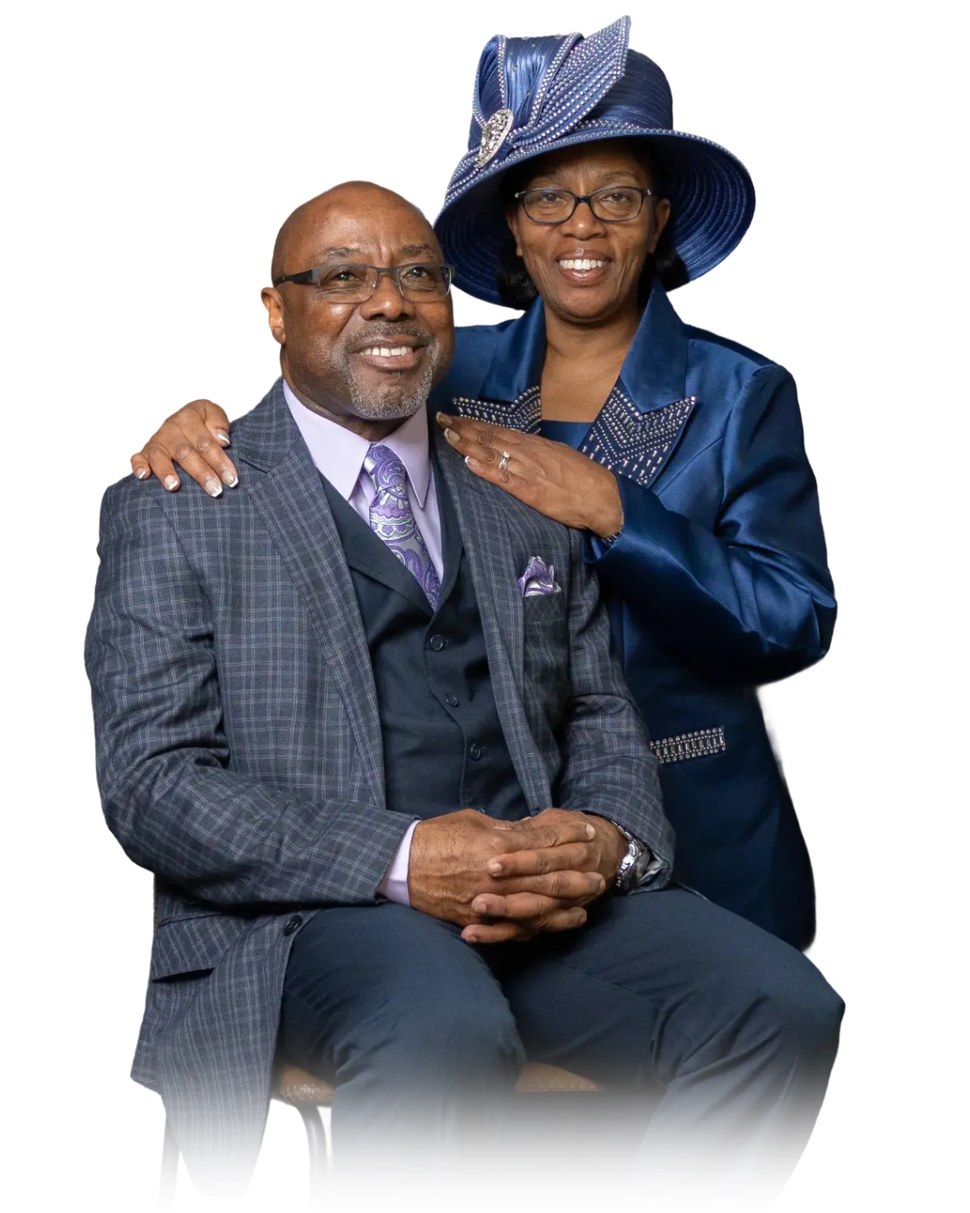 A smiling man in a plaid suit and glasses sitting on a chair, with a woman in a blue outfit and large matching hat standing behind him with her hand on his shoulder, against a black background.