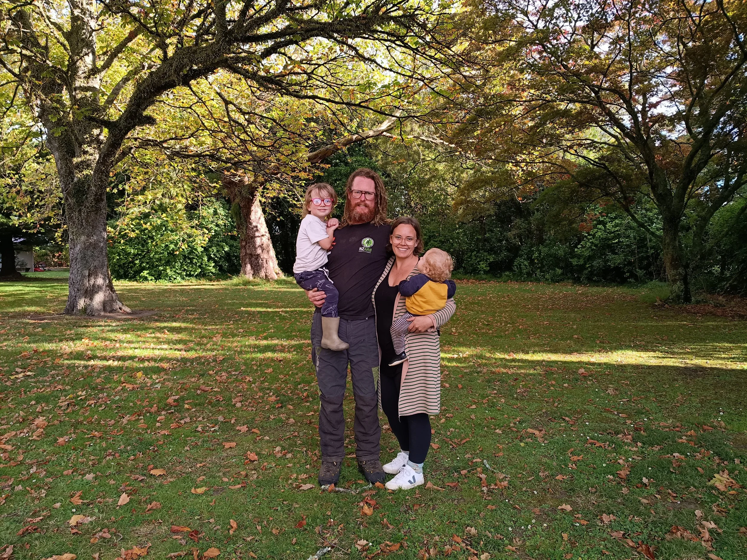 A family photo of four standing in a park with trees and fallen leaves. The man is holding a young boy and a woman is holding a toddler. All are smiling.