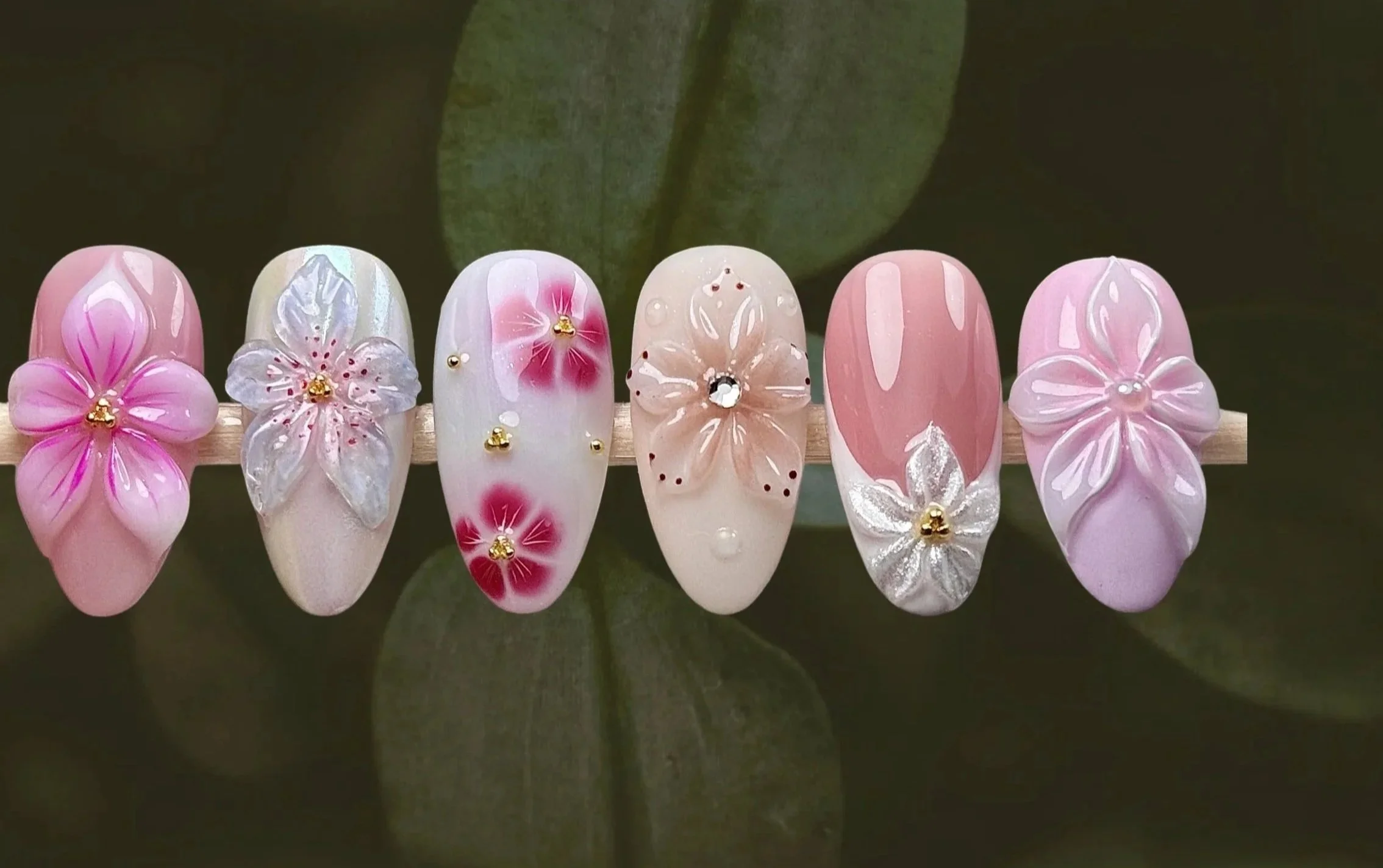 Serie de uñas acrílicas decoradas con diseños de flores en tonos rosa, blanco y con detalles en dorado y brillante.