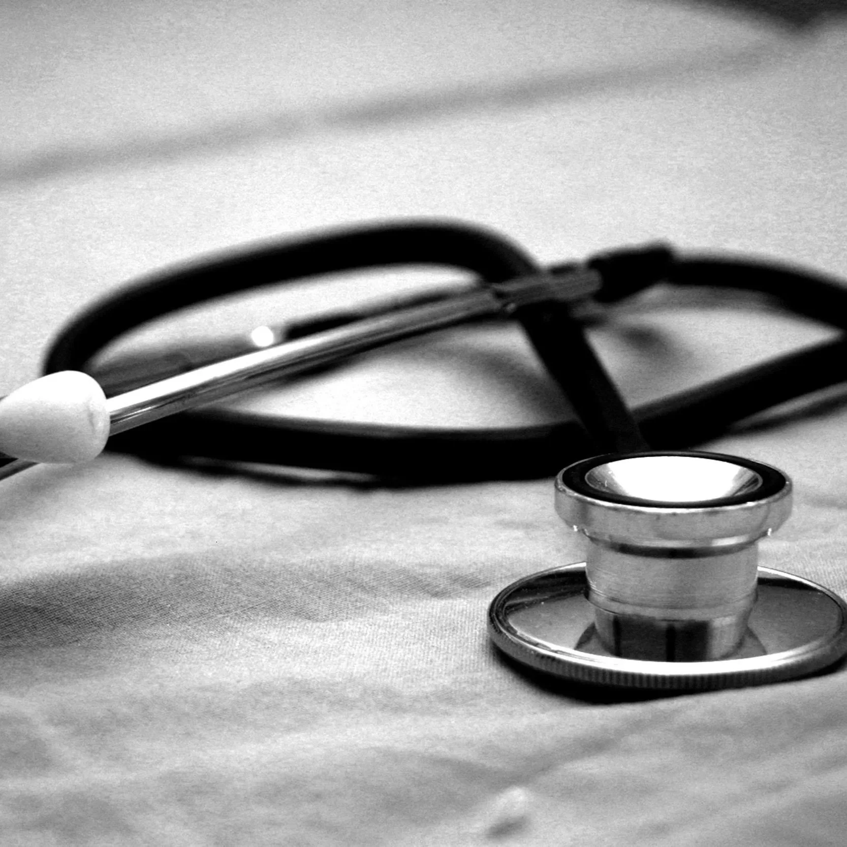 Black and white close-up of a stethoscope placed on a surface, with the chest piece in focus and the tubing coiled in the background.