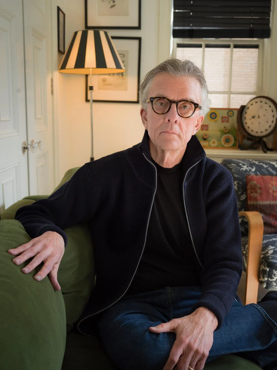 Kurt Andersen. An older man with gray hair, wearing black glasses, a black zip-up sweater, and a black shirt, sitting on a green couch in a cozy, well-decorated living room.