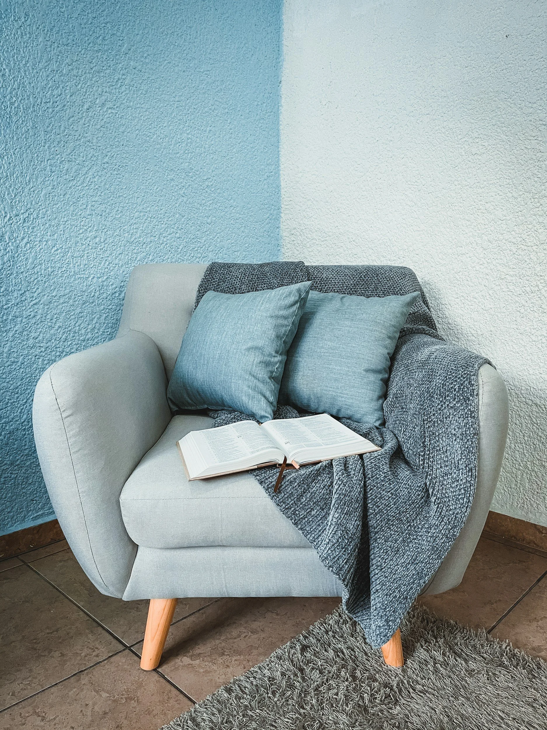 Picture of a light grey, comfortable arm chair. On the armchair are two blue cushions, an open book, and there is a grey textured throw draped over the arm.