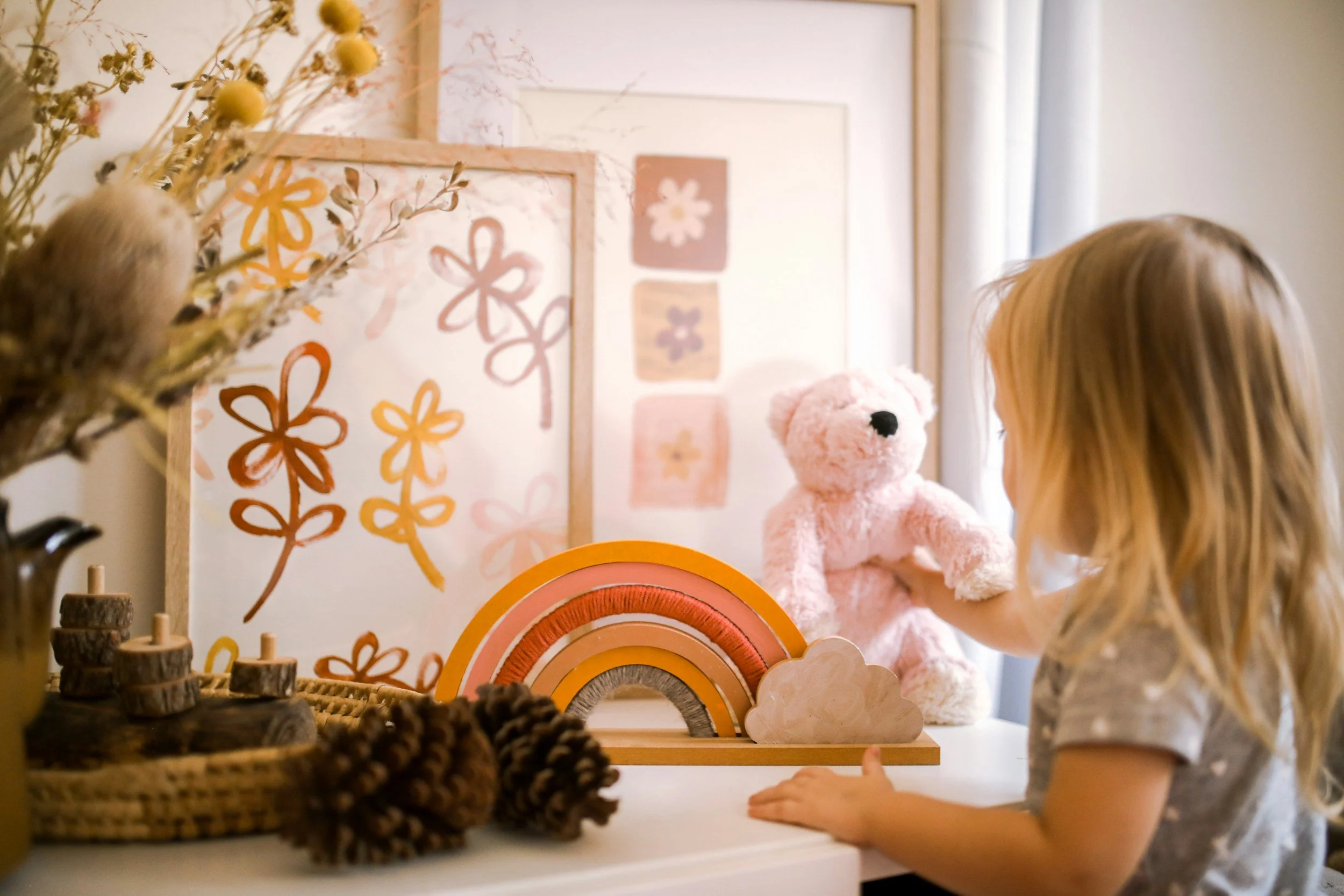 Picture of a child playing with a teddy bear in a room decorated with drawings of flowers. On the table next to the child are pinecones and a rainbow.