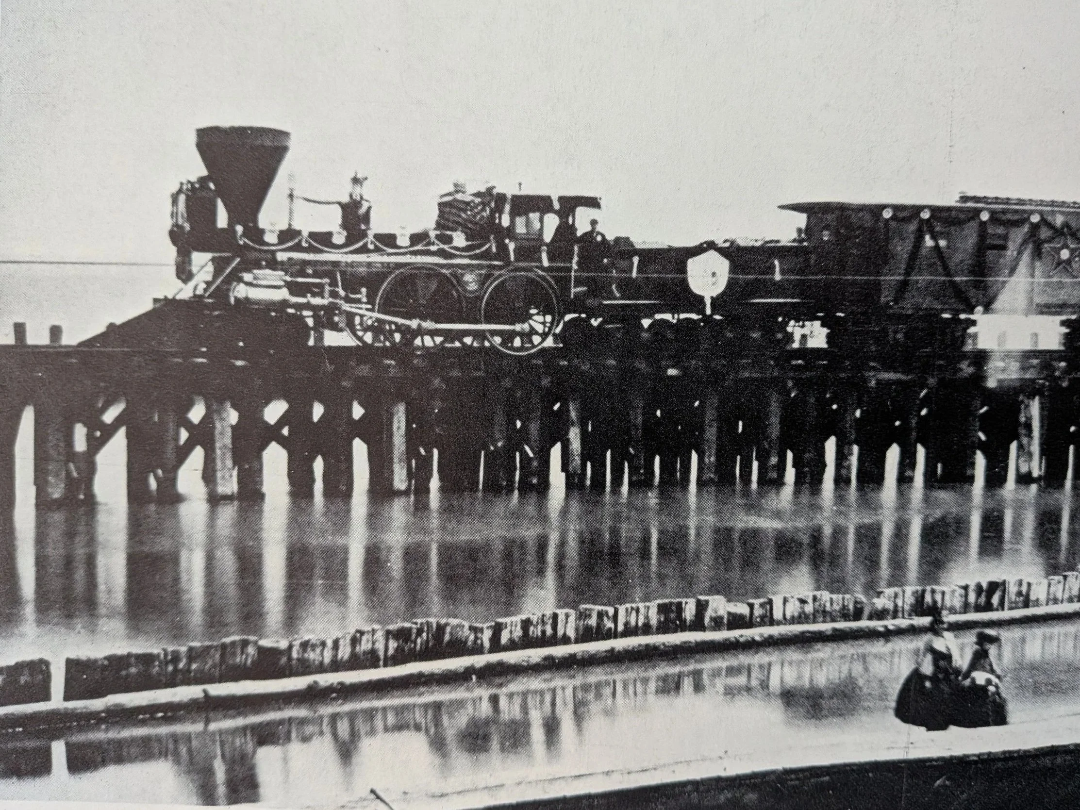 A black and white photo of a vintage steam locomotive on a pier over water, with a person riding a motorcycle in the foreground.