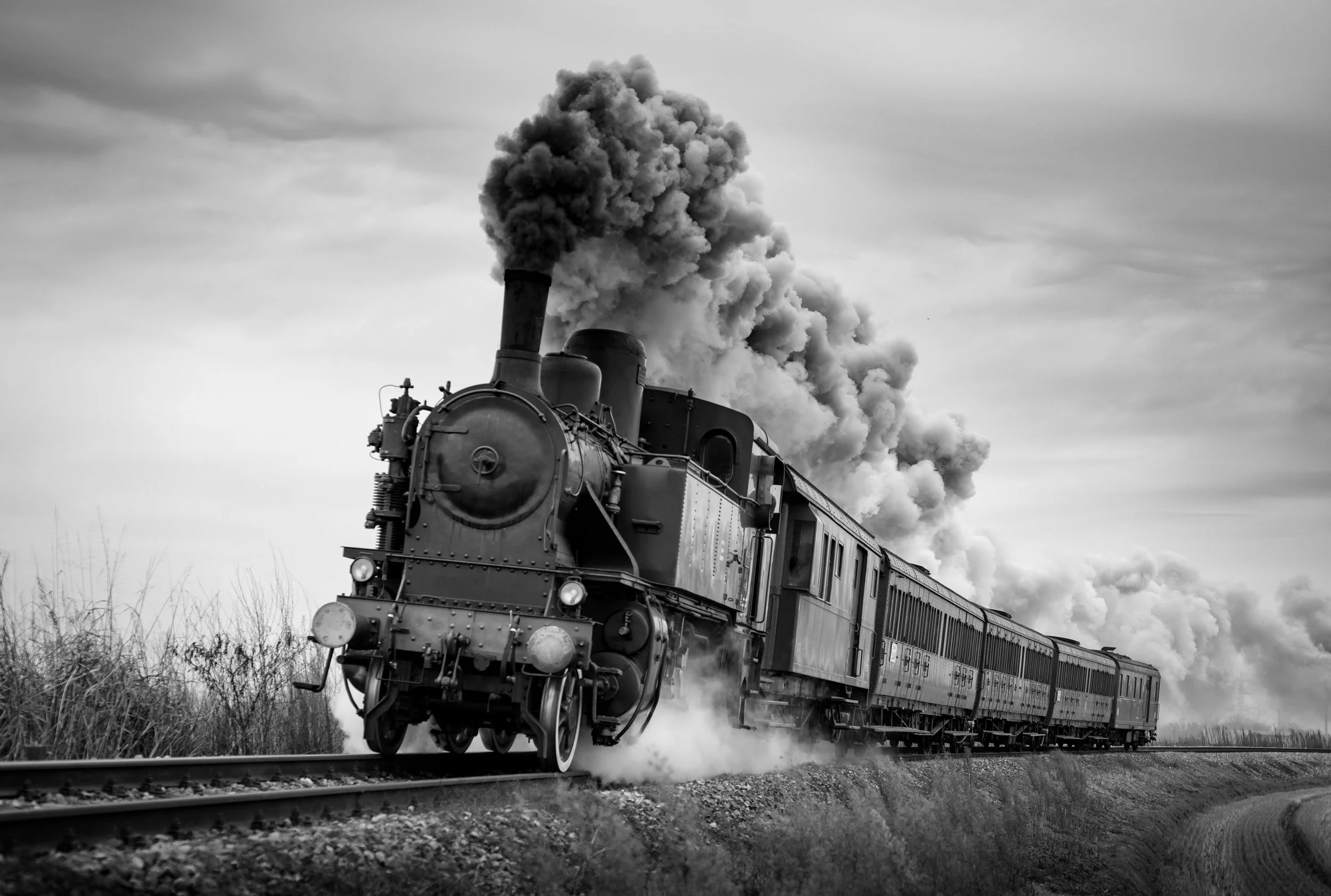 A black and white photograph of a vintage steam locomotive train emitting large clouds of smoke as it travels along a track through a rural landscape.