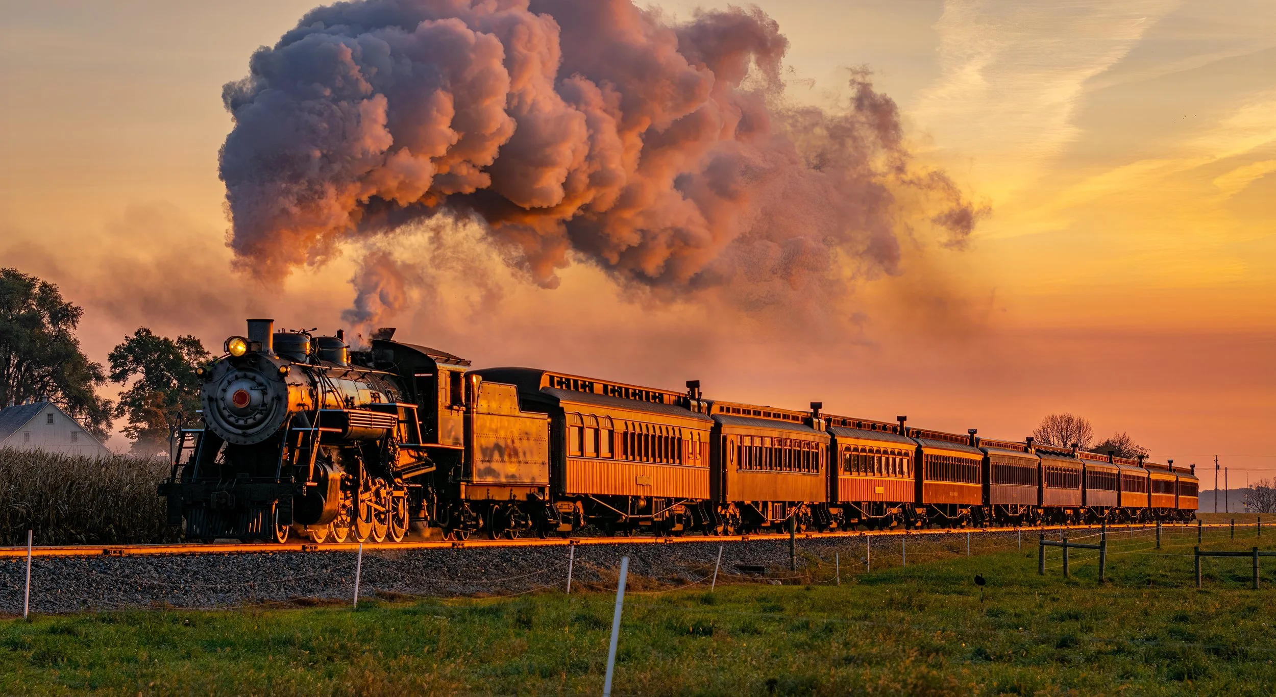 A vintage steam train with multiple cars traveling through a rural landscape at sunset, with smoke billowing from the smokestack and an orange glow reflecting on the train and sky.