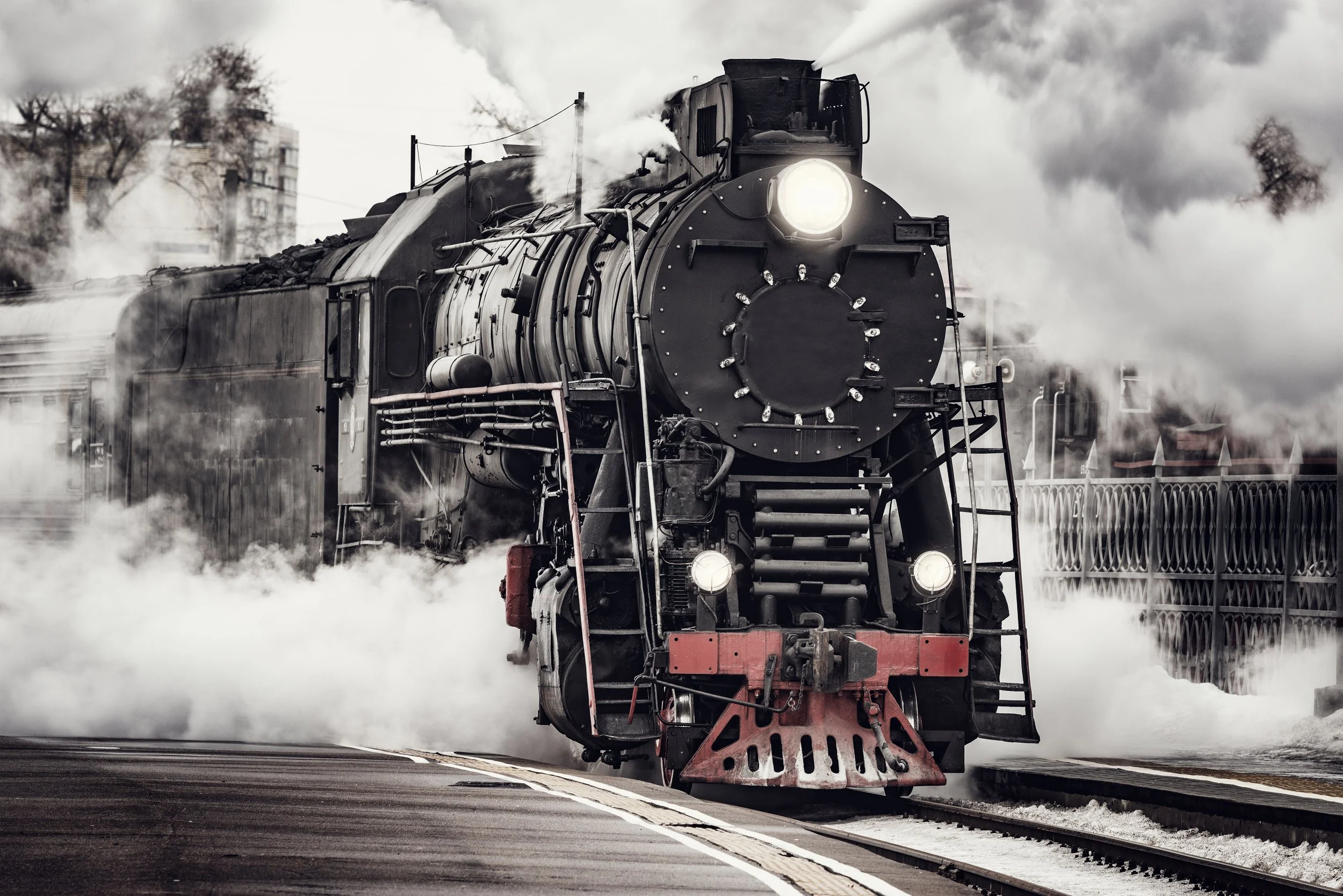 Black steam locomotive emitting steam and smoke, approaching a railway station platform on a cloudy day, with trees and buildings in the background.