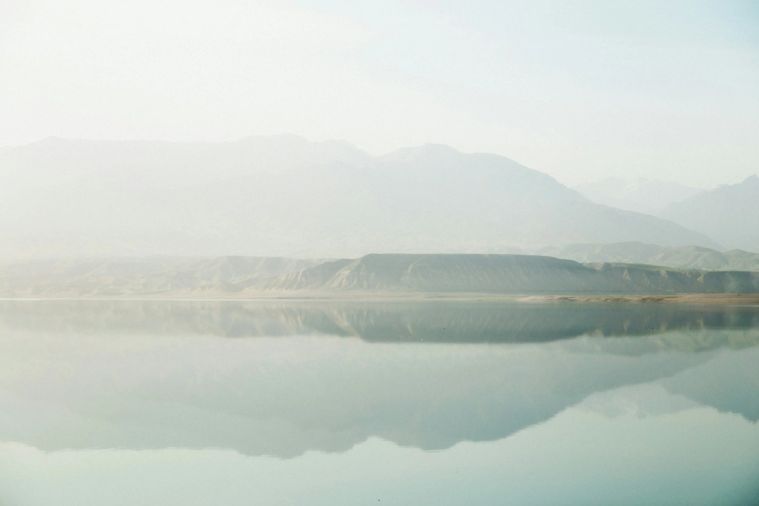 A calm lake reflecting distant mountains and hills under a light, hazy sky.