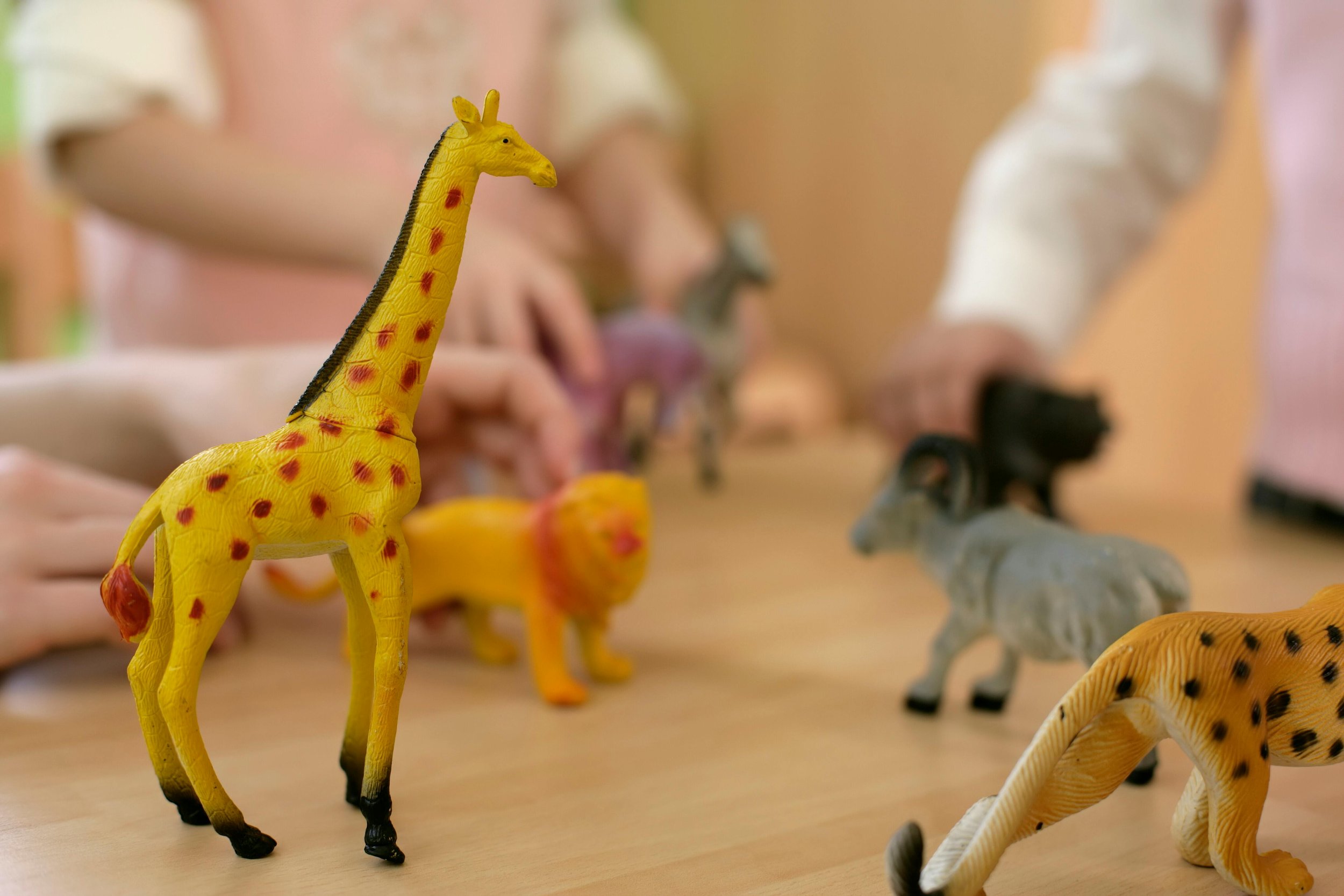 Colorful toy animal figures, including a yellow giraffe with red spots, on a wooden table. In the background, adolescent in play therapy.