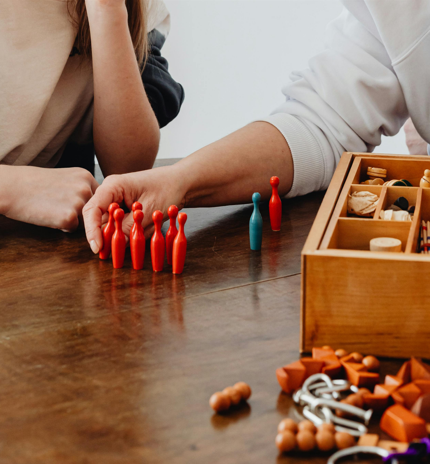 Two people playing a tabletop game with red and blue game pieces in front of a wooden storage box filled with game accessories. Play therapy.