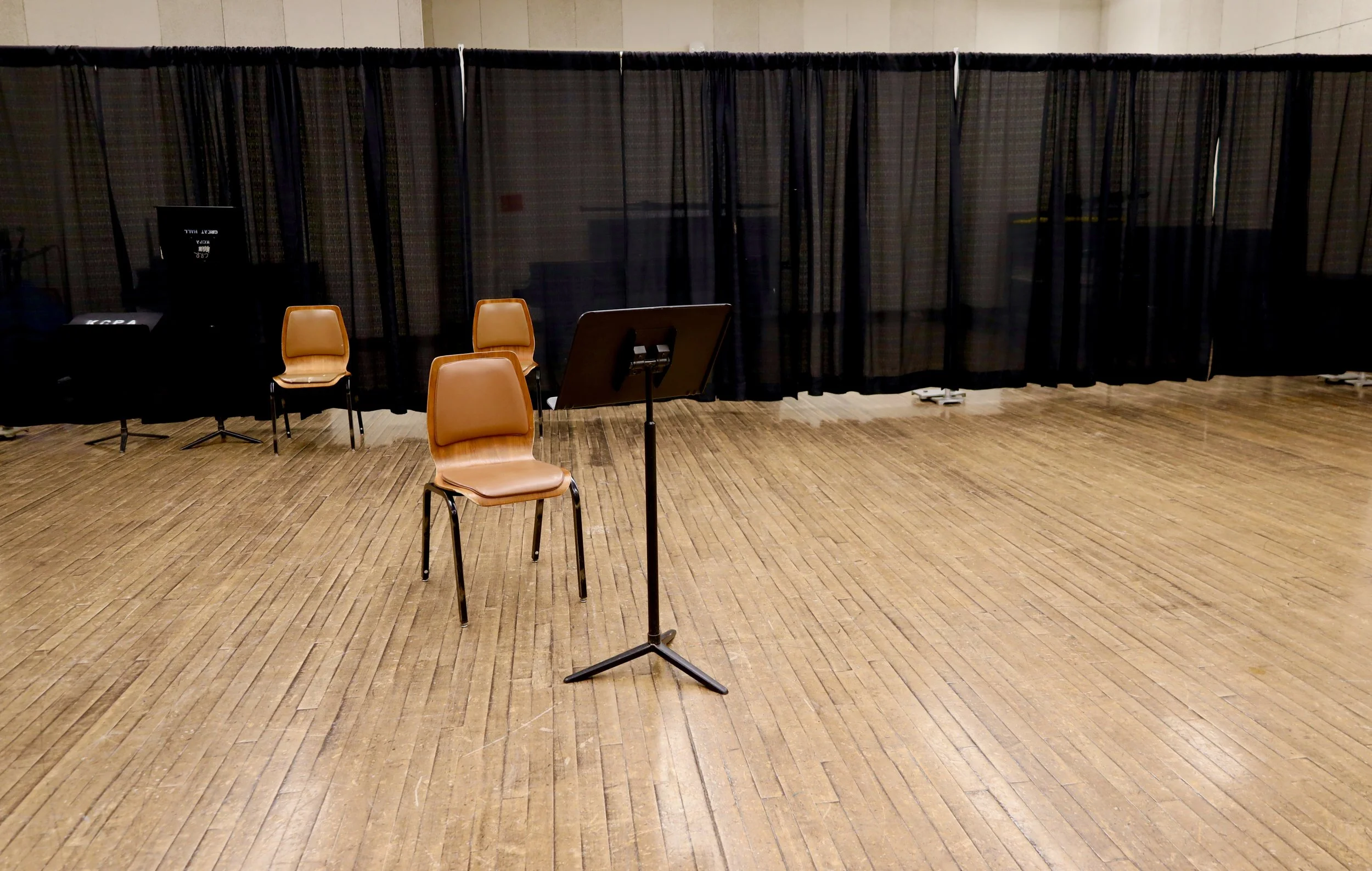 Empty stage with a black curtain backdrop, a music stand at the front, three chairs, and some stage equipment.