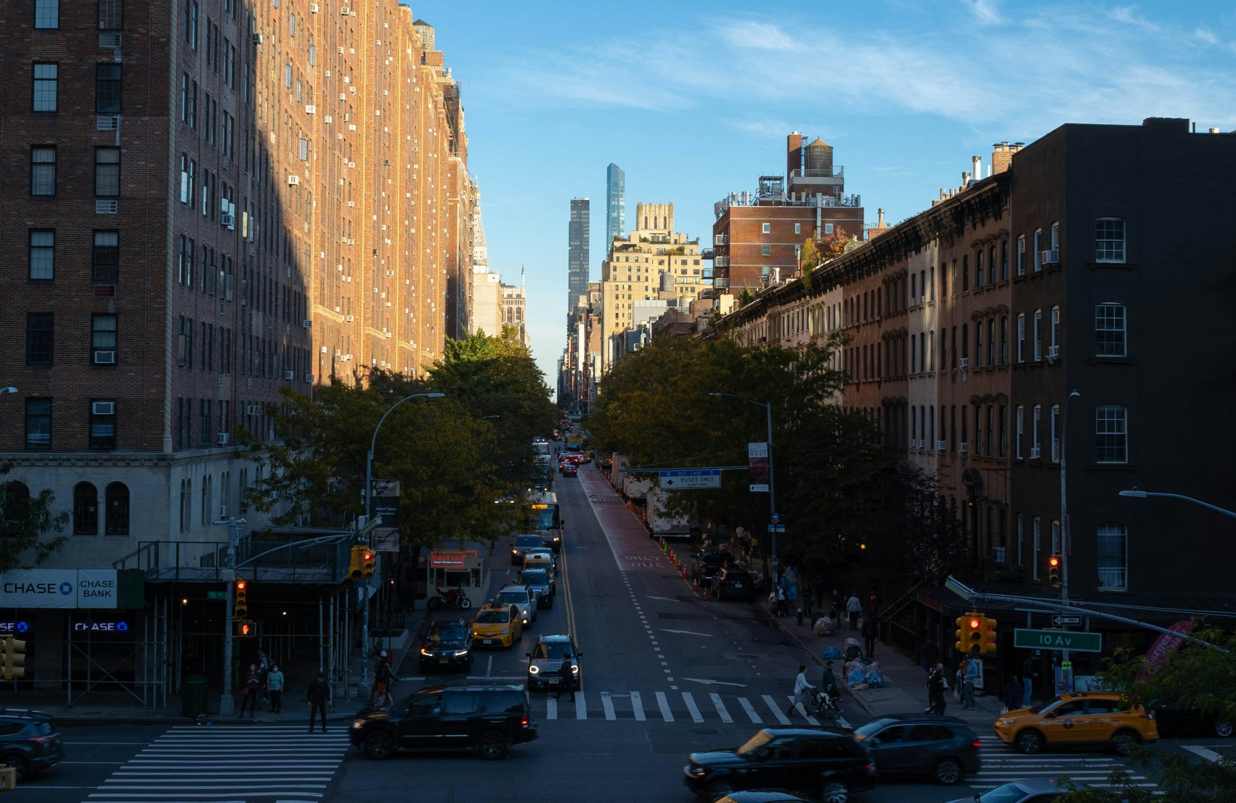 New York City neighborhood with tall buildings on both sides, cars and taxis on the road, and pedestrians crossing at the crosswalk in late afternoon sunlight.