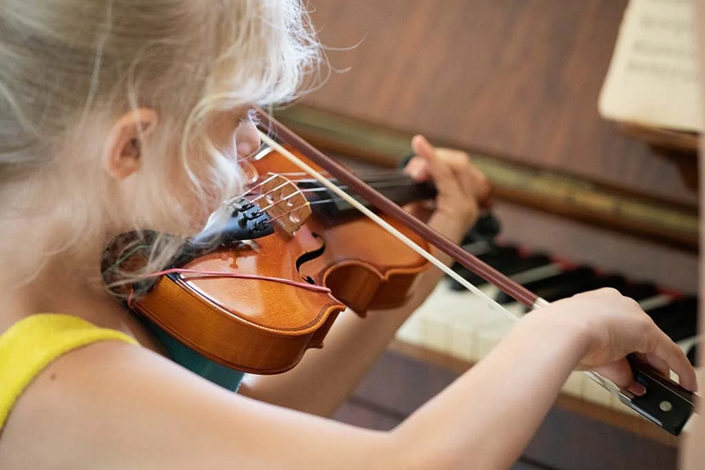 A young girl playing the violin while accompanied by a piano.