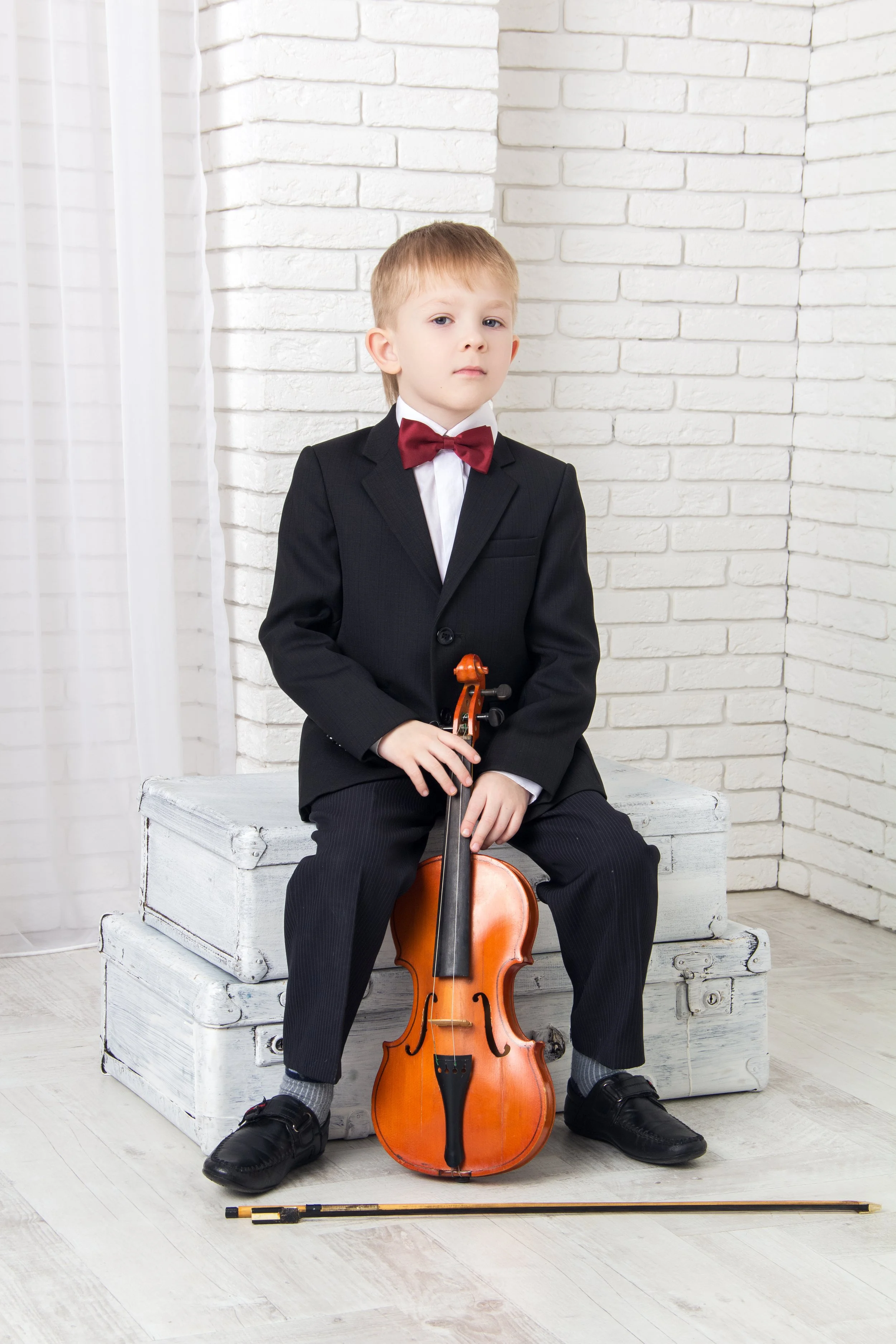 A young violin student in a formal black suit with a maroon bow tie is sitting on a white wooden chest, holding a violin.