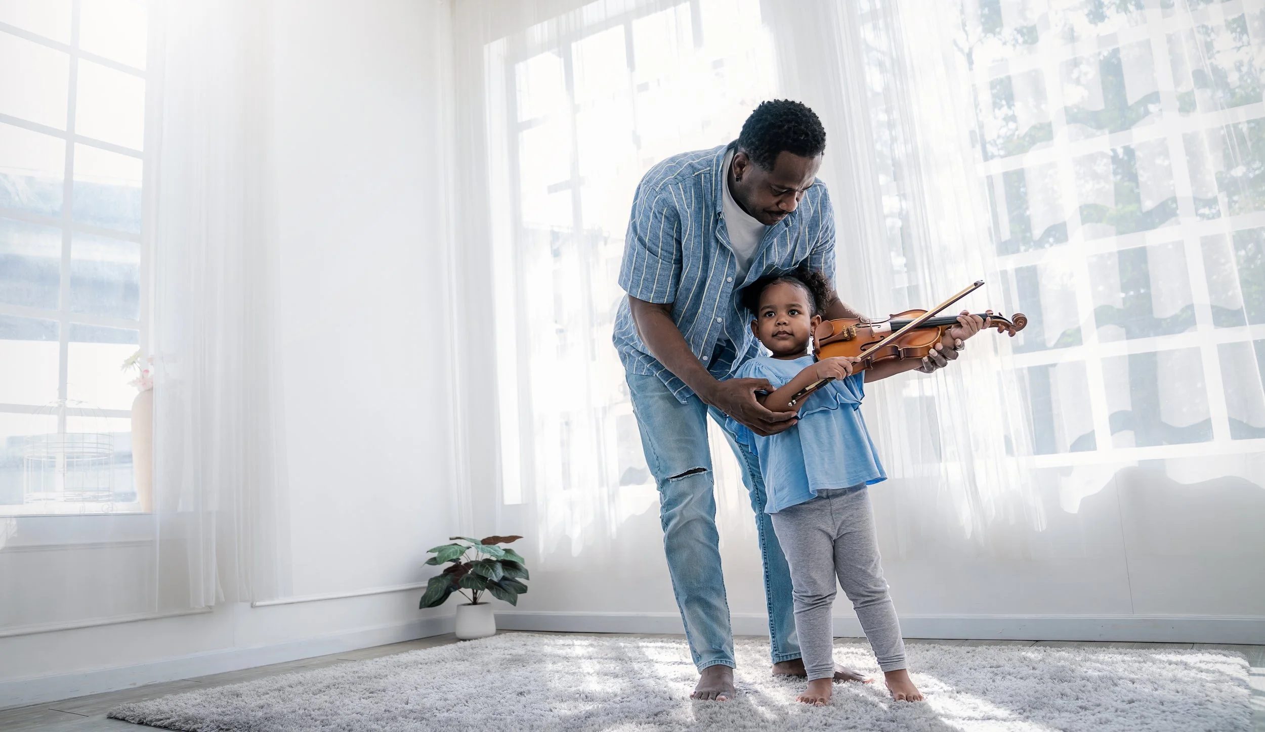 A parent helping his young daughter hold a violin inside a bright living room with large windows and white curtains.