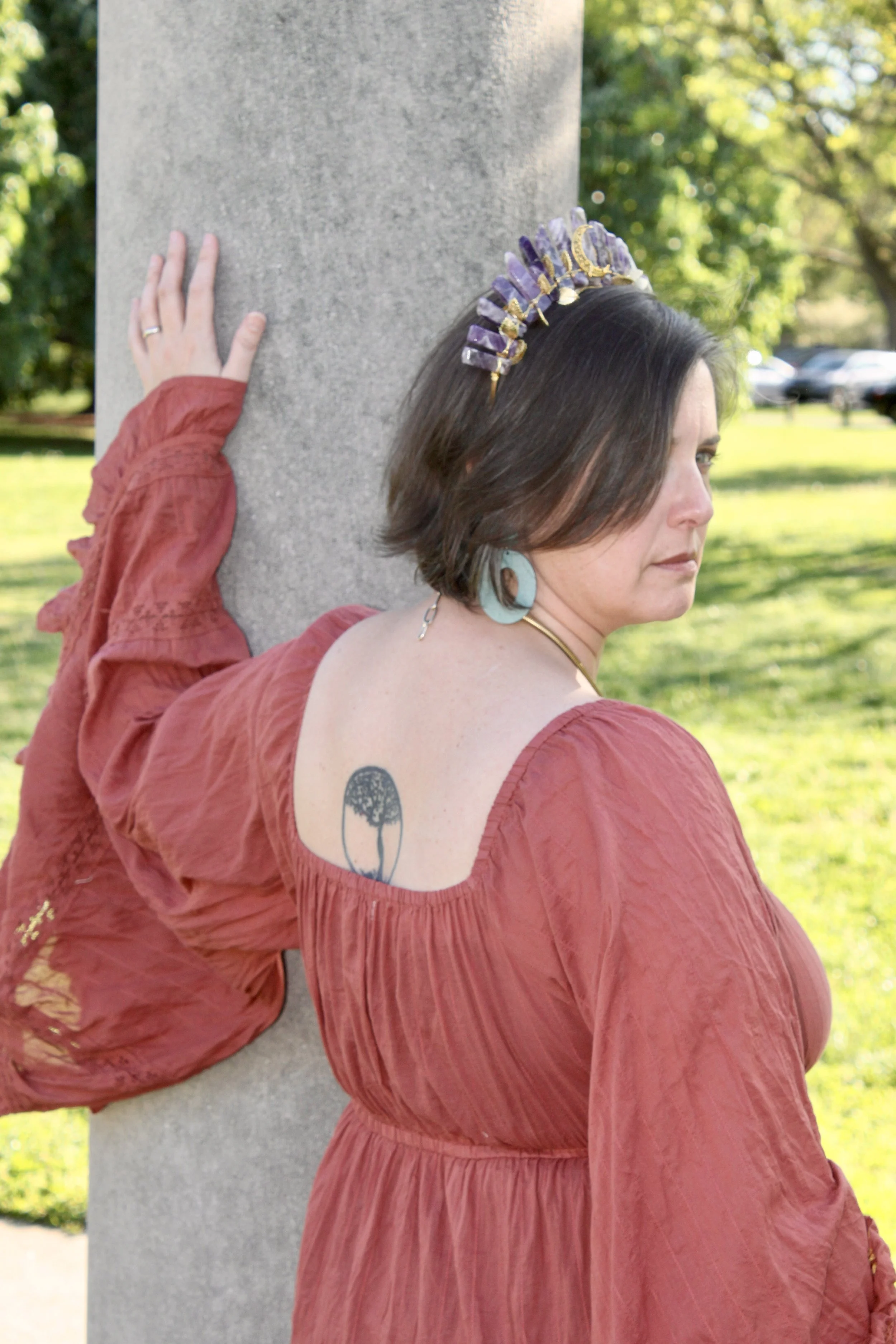 A woman with short dark hair, wearing large turquoise earrings, a deranged headband, and a reddish-brown dress with long sleeves, is depicted standing outdoors in front of a concrete column, with her hand resting on the column. There are trees and parked cars in the background.
